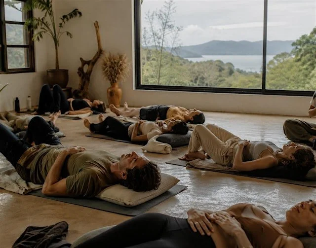 Group of people practicing yoga or meditating indoors on mats near a large window with a view of trees and water.