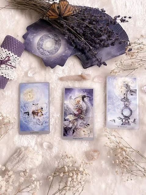 Tarot cards with mystical artwork spread out on a soft, light-colored fabric, accompanied by dried lavender, a lace cloth, and decorative elements setting a mystical, dreamy scene.
