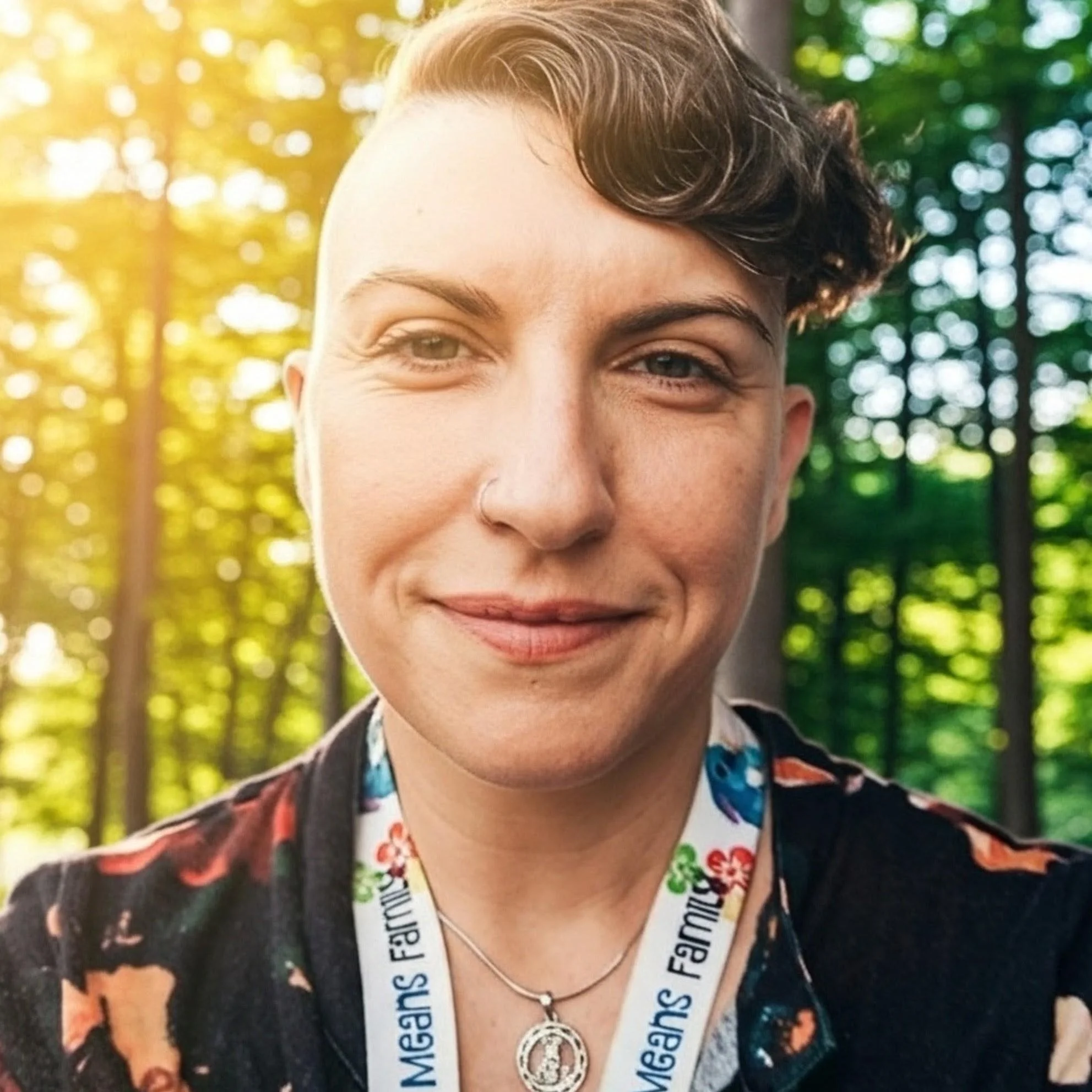 Close-up of a smiling person with short, curly hair outdoors with greenery in the background, wearing a patterned shirt and a necklace.