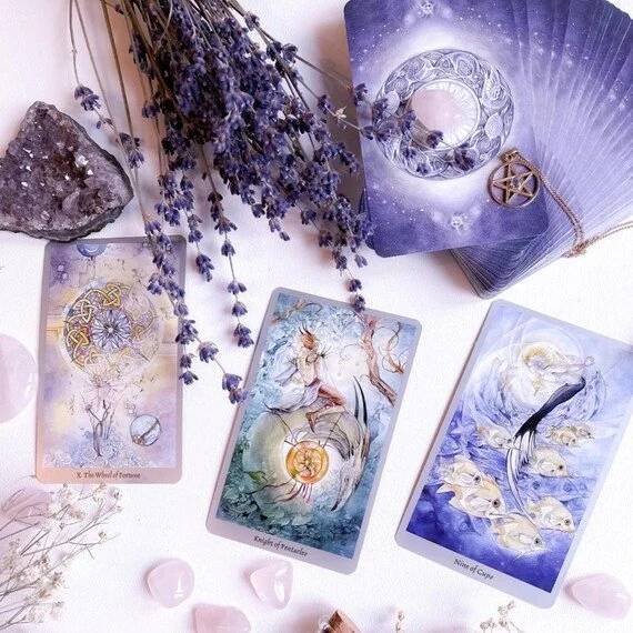A tarot card reading setup with three tarot cards, a bundle of lavender, a large crystal, pink stones, and a decorated fan with lunar and pentagram symbols.