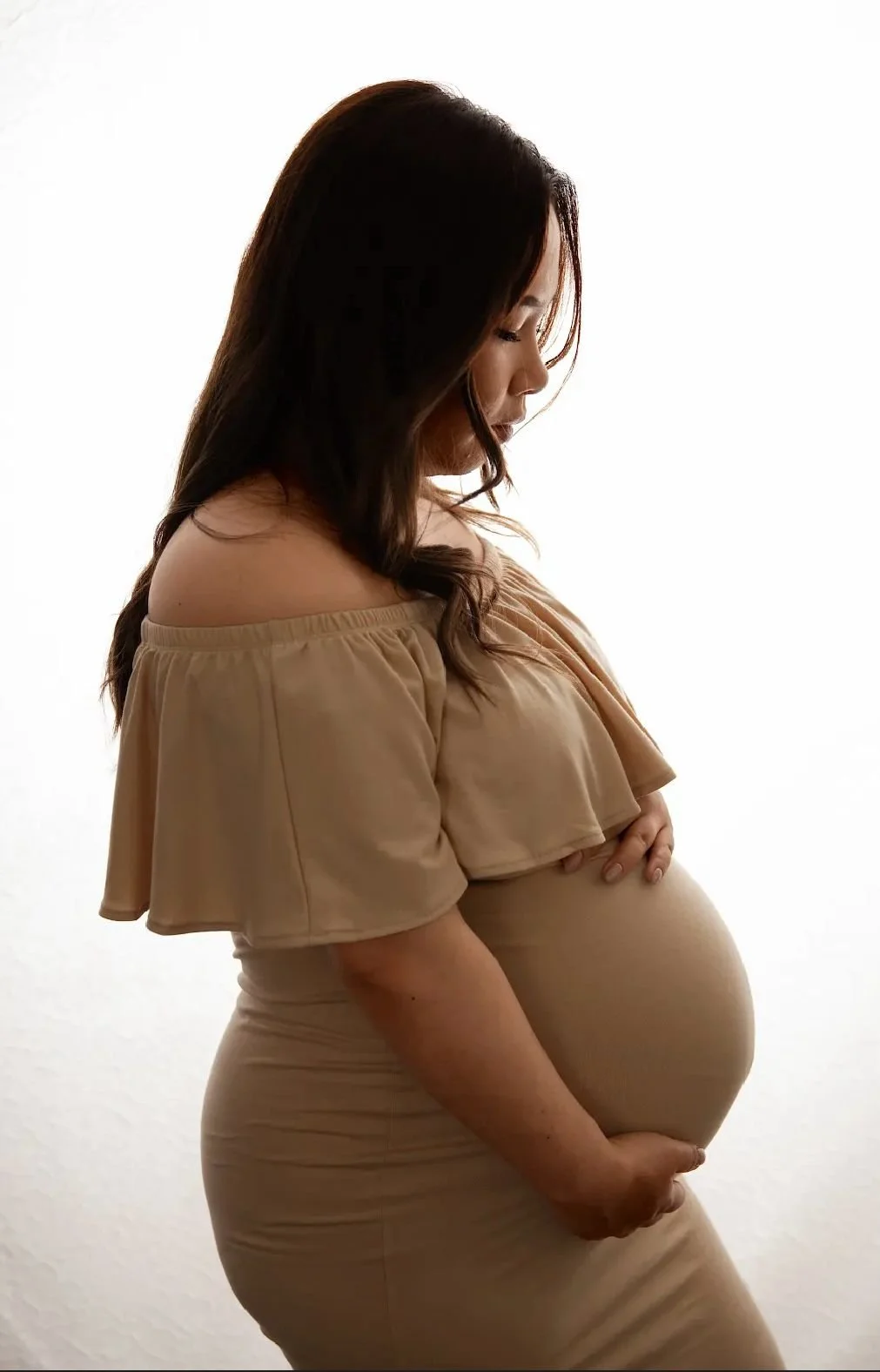 A pregnant woman with long dark hair in a beige off-the-shoulder dress stands sideways against a white background, cradling her belly with one hand.