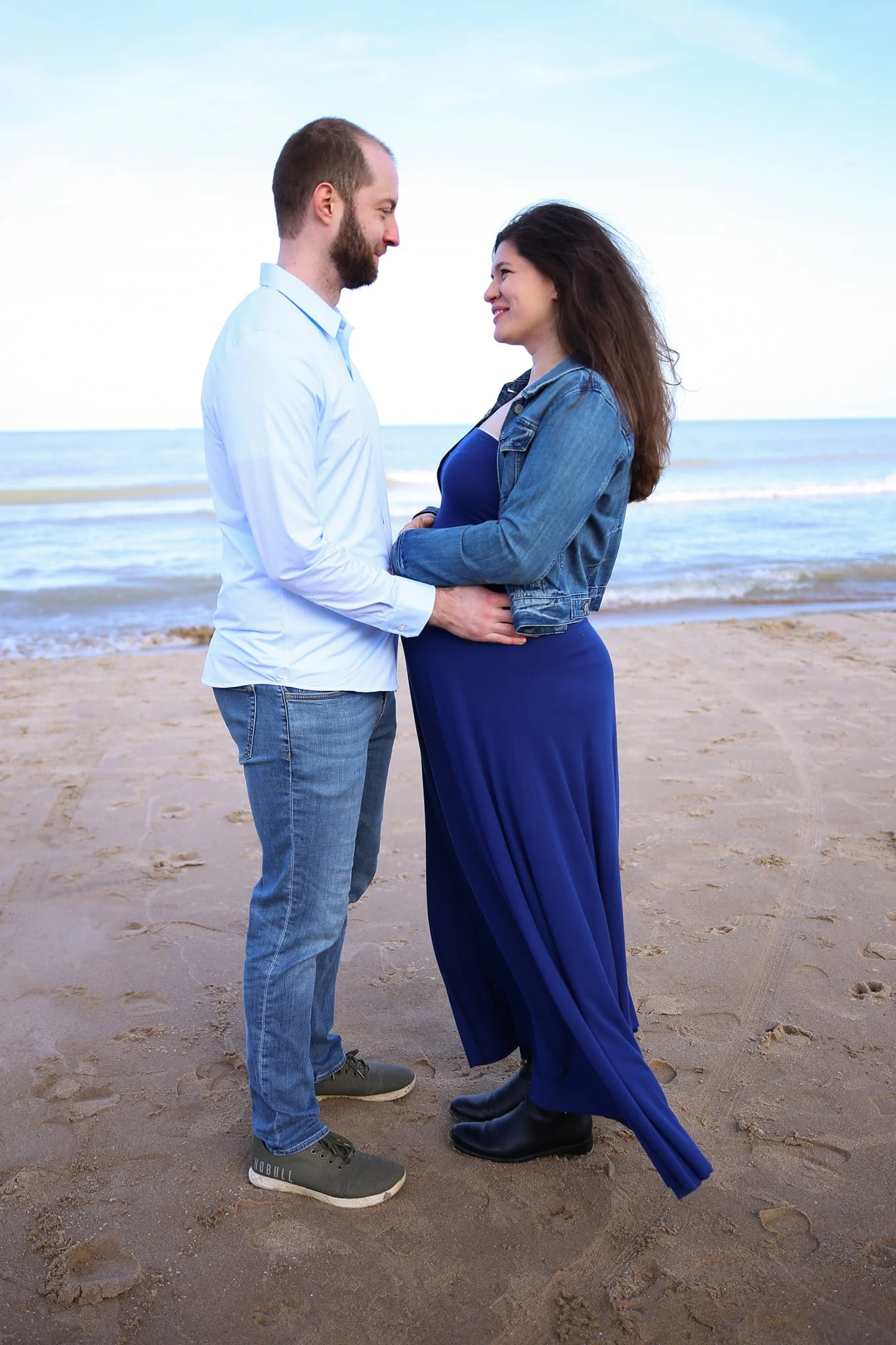 A couple standing close together on a beach facing each other, smiling, with the ocean in the background.