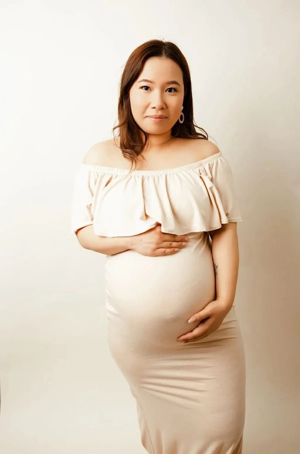 A pregnant woman in a beige off-the-shoulder dress holding her belly and touching her chest, standing against a plain background.