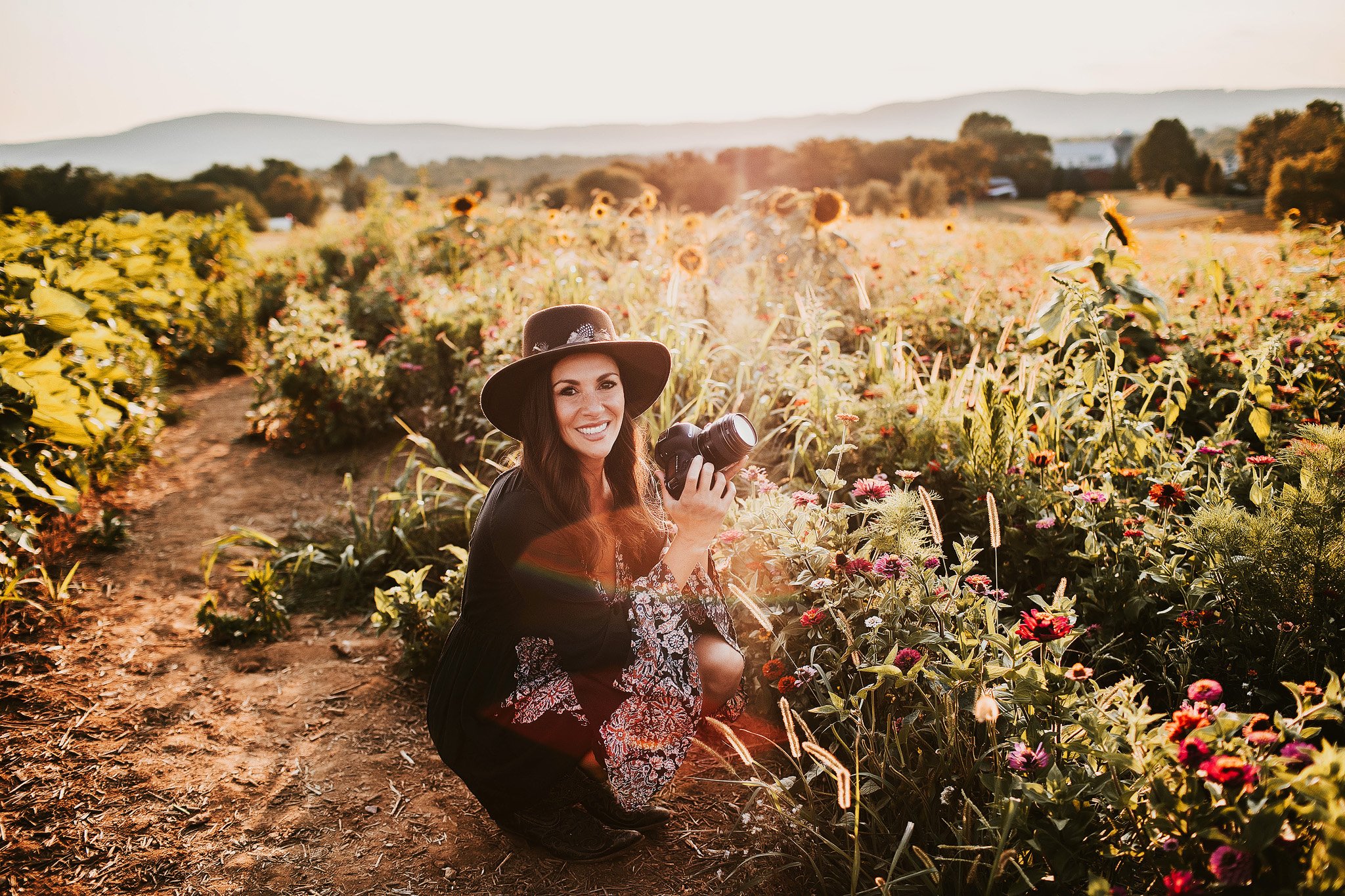 A woman with a wide-brimmed hat kneeling on a dirt path holding a camera in a sunflower and flower field at sunset.