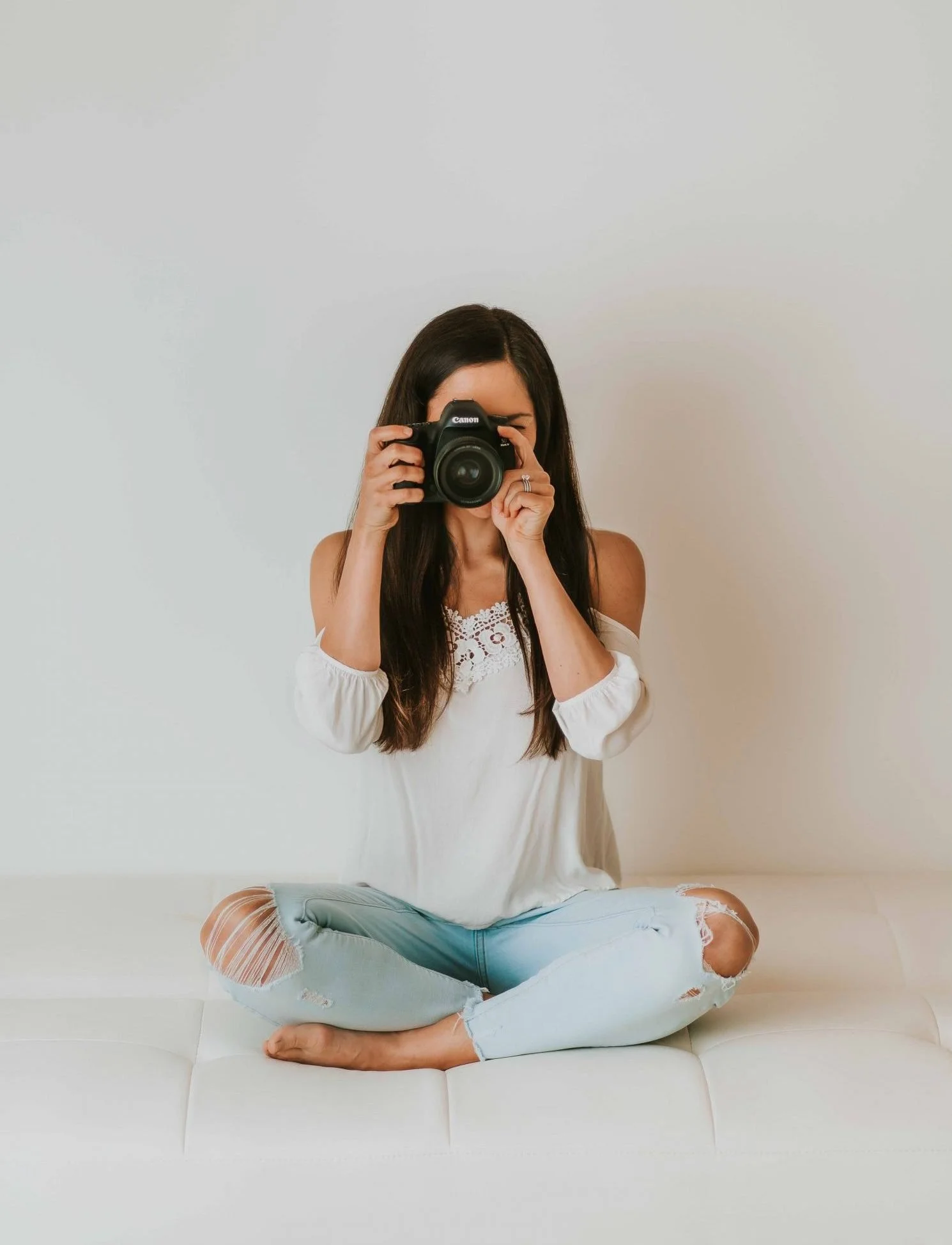 Young woman sitting cross-legged on a white bench, holding a camera up to her face and taking a photo, against a plain white wall.