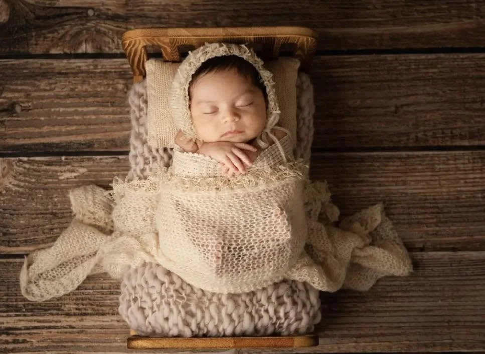 Photo of a sleeping baby wrapped in a cream-colored knitted blanket, wearing a matching bonnet, lying on a small wooden bed on a wooden floor.