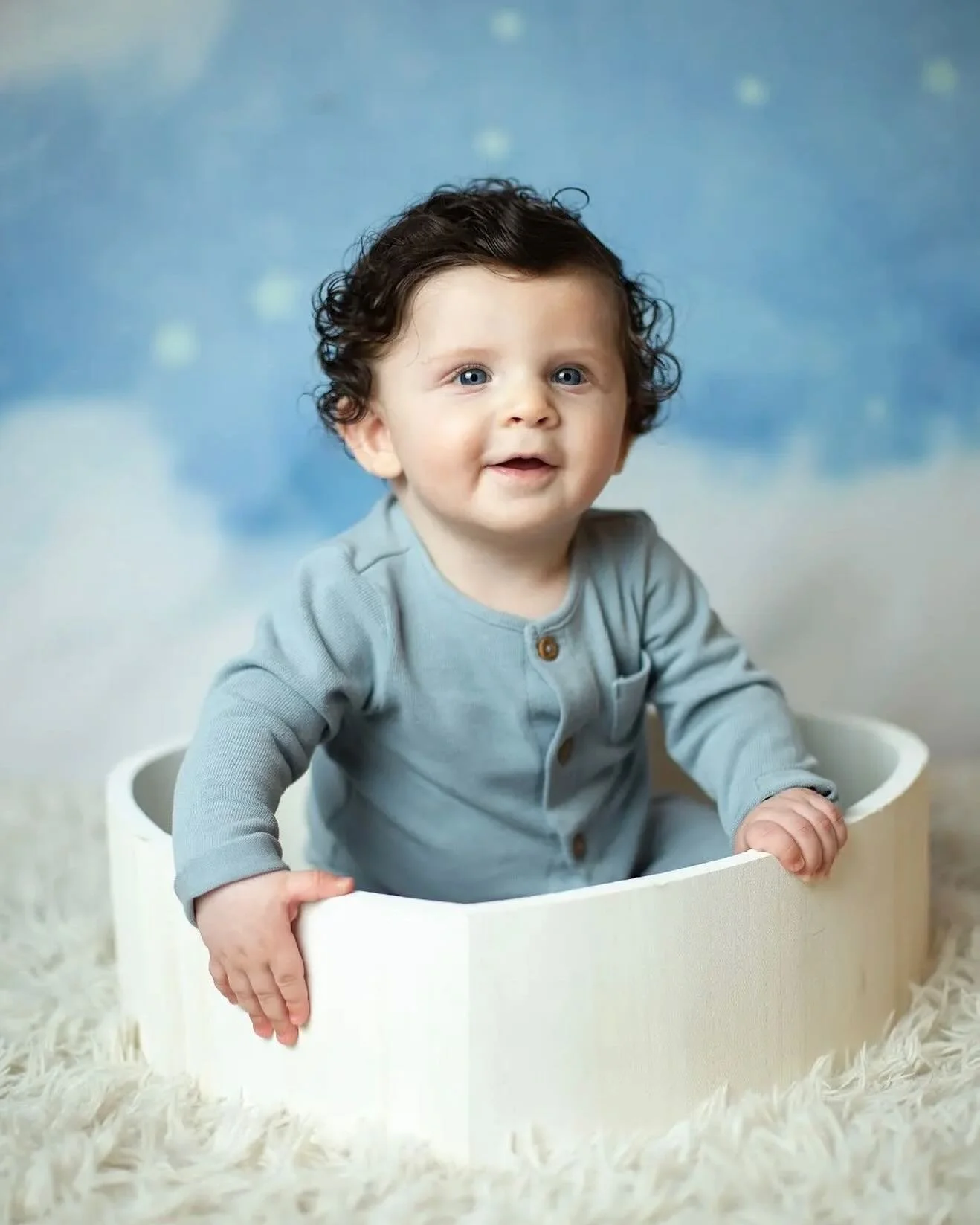 Young child with curly dark hair and blue eyes sitting in a white box on a fluffy cream-colored rug, wearing a light blue long-sleeve shirt, with a blue blurred background.