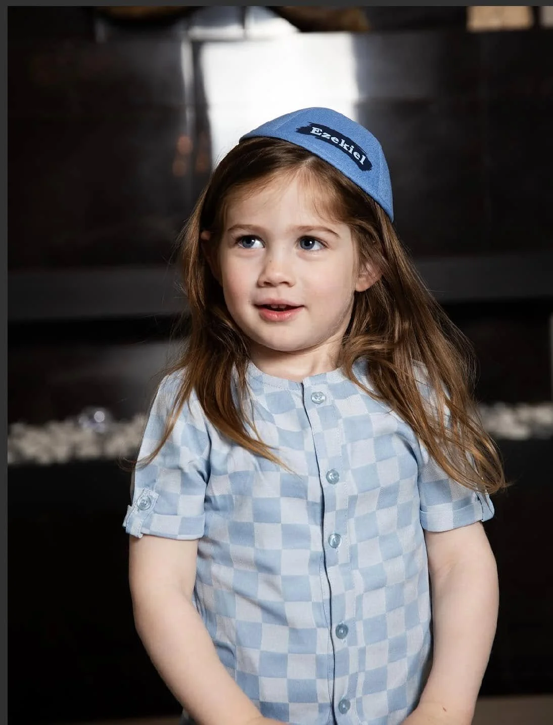A young girl with long red hair and blue eyes wearing a blue kippah with Hebrew writing and a light blue checkered shirt, standing indoors.