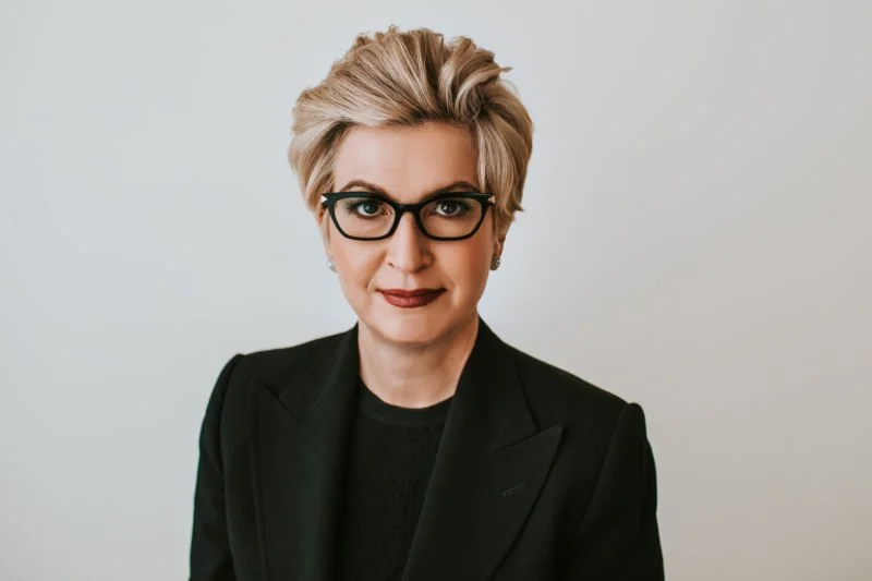 A woman with short blonde hair, wearing black glasses and a black blazer, standing against a plain white background.