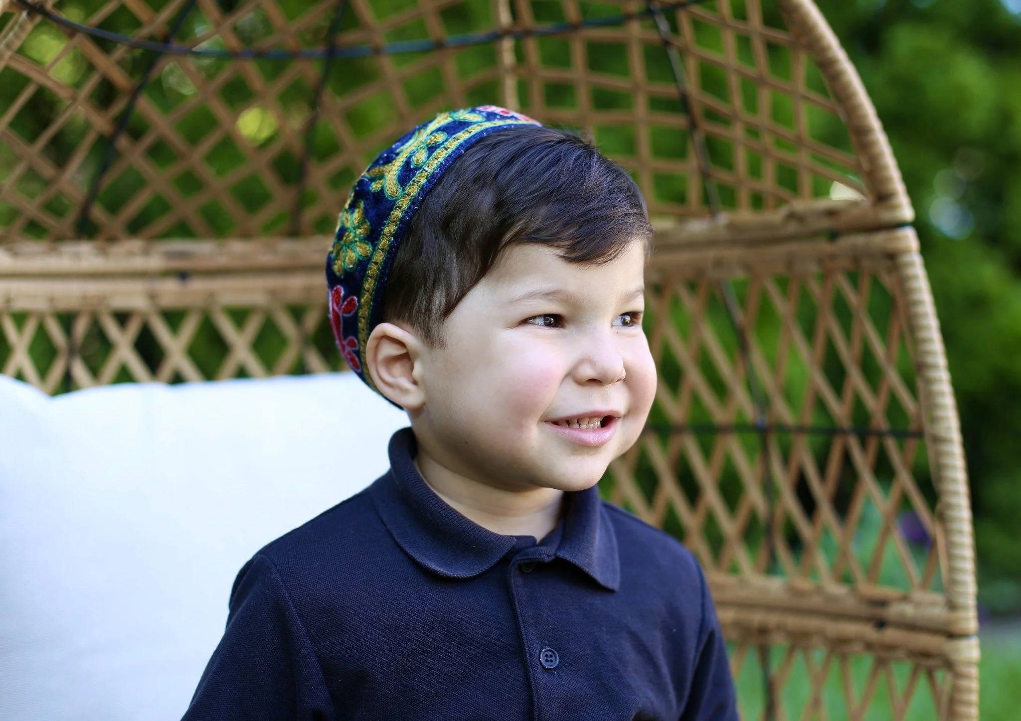 A young boy with dark hair, wearing a navy blue shirt and a colorful embroidered cap, sitting outdoors in front of a woven wicker structure and a white cushion.