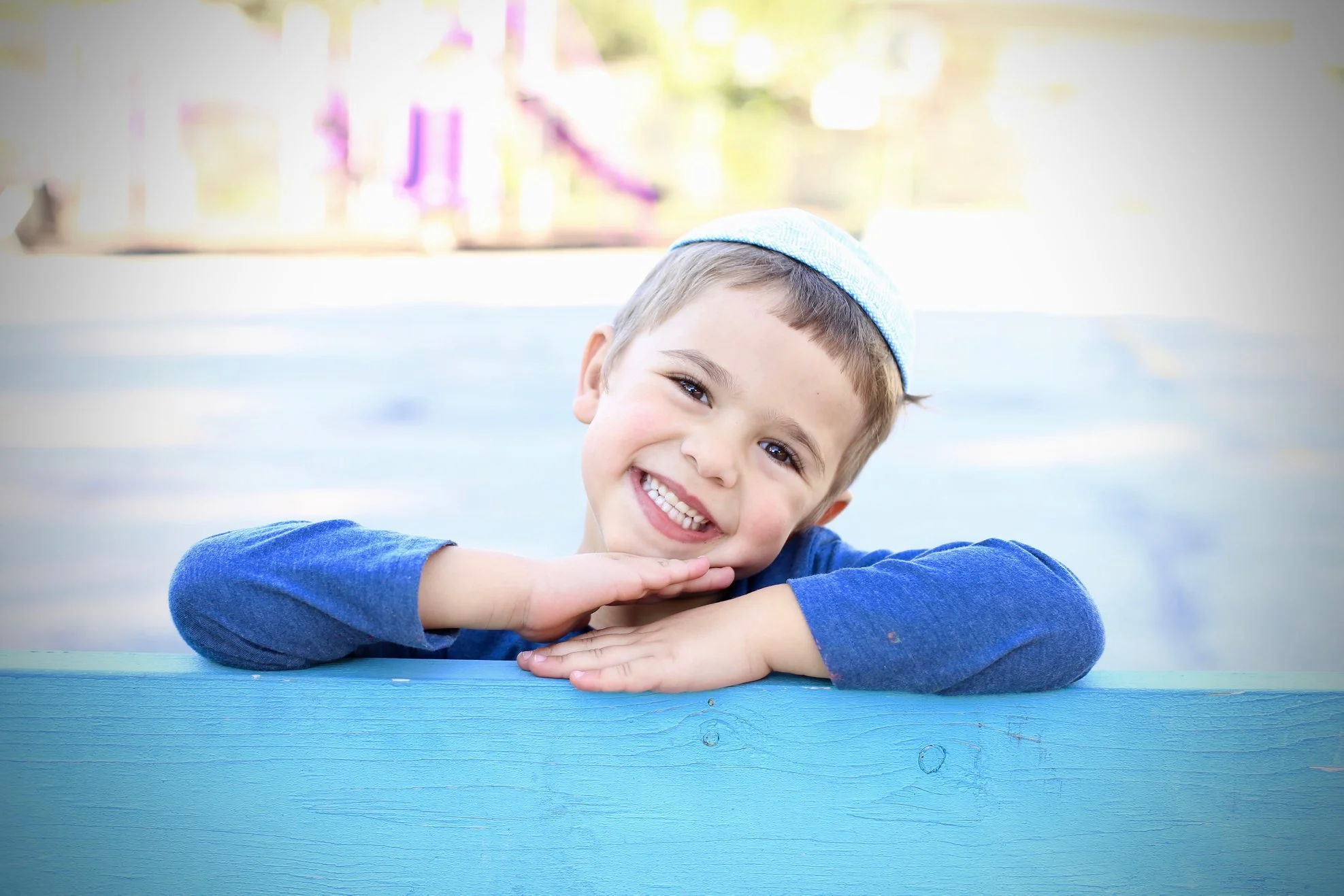 A young boy with a big smile, wearing a blue hoodie and a blue kippah, resting his head on his hands on a blue surface outdoors.