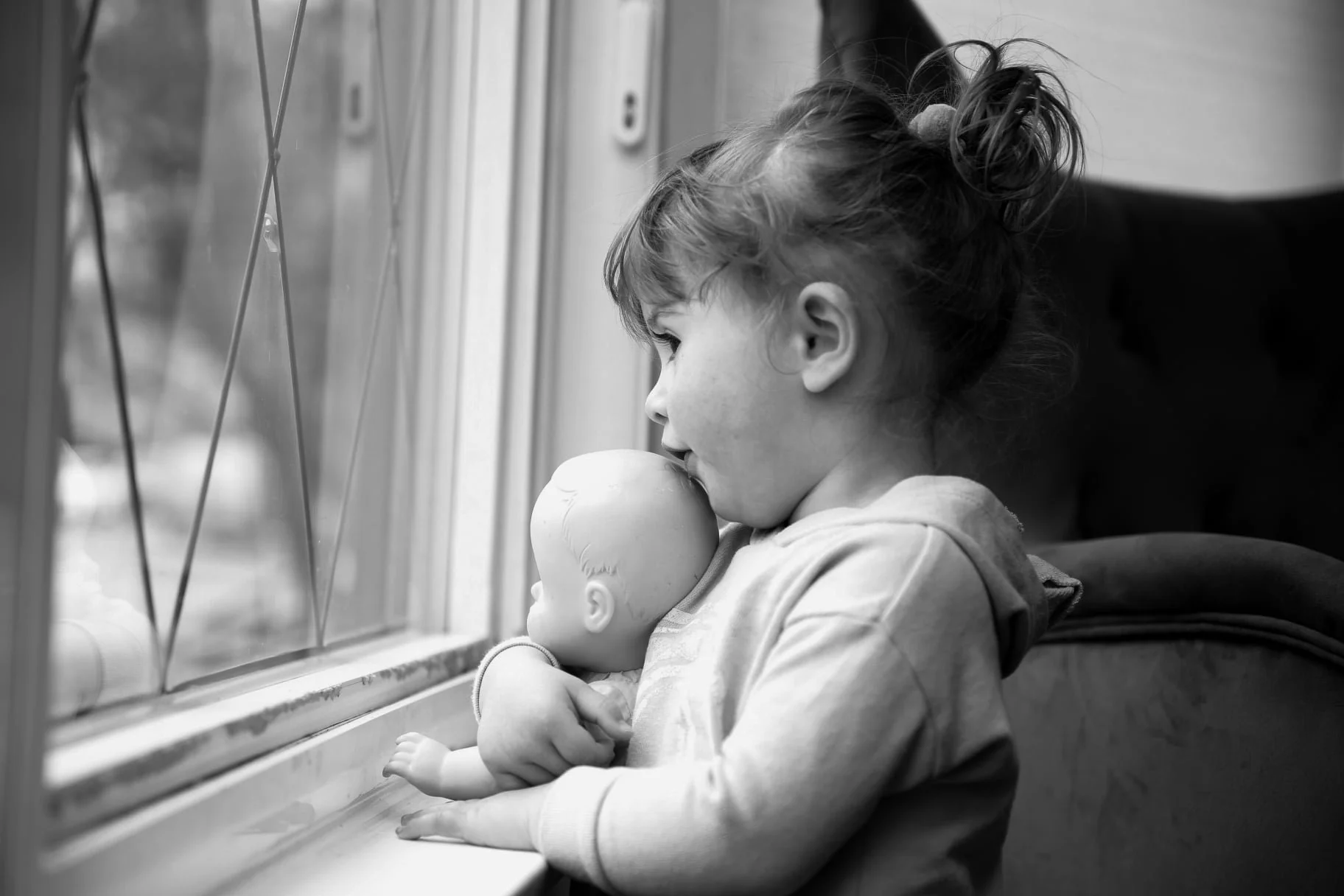 A young girl with her hair tied up in a messy bun, gazing out the window, holding a doll while sitting on a seat.
