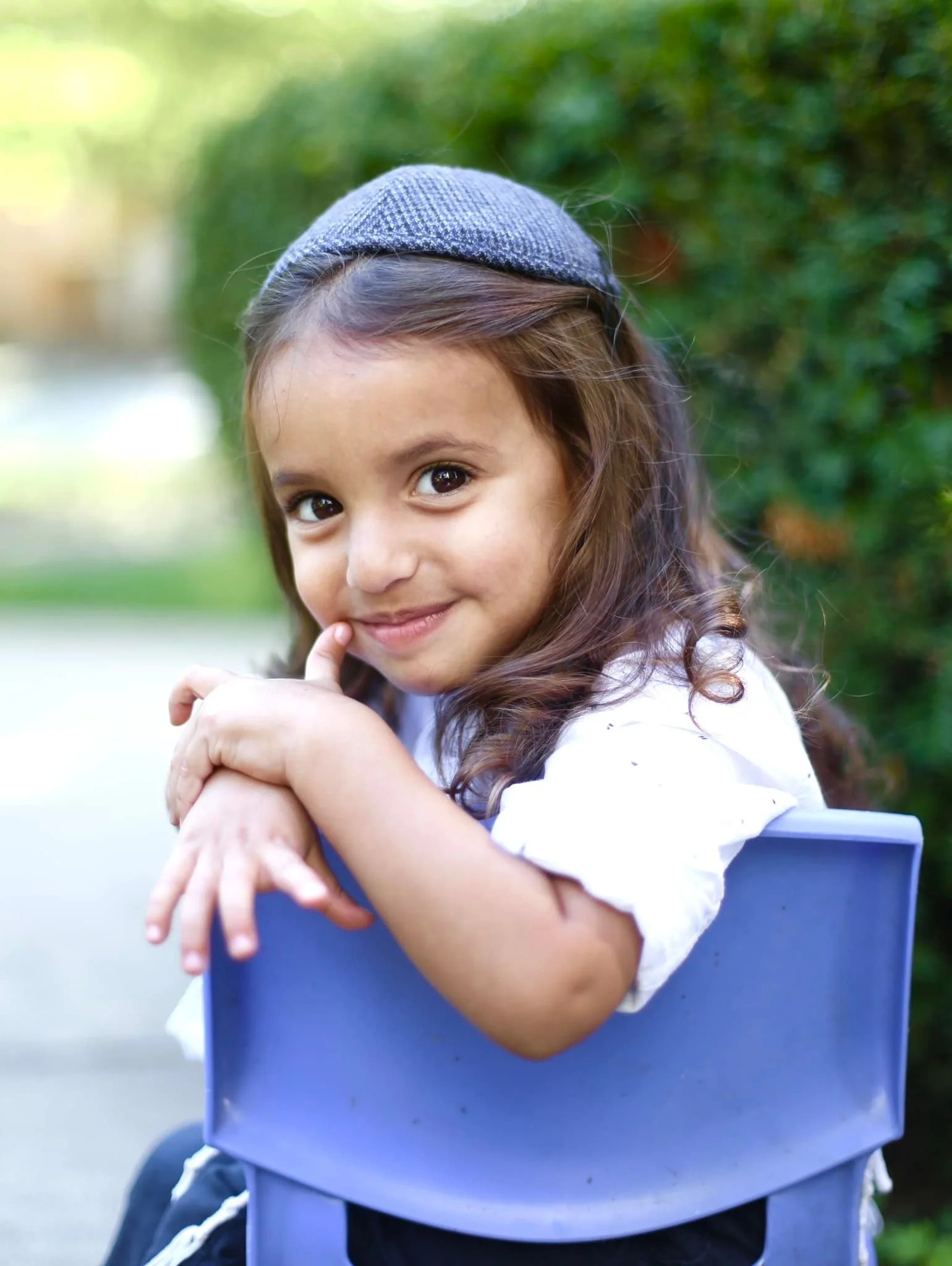 A young girl with brown hair wearing a gray knit hat, sitting on a blue chair outdoors, smiling and resting her chin on her hands, with lush greenery in the background.