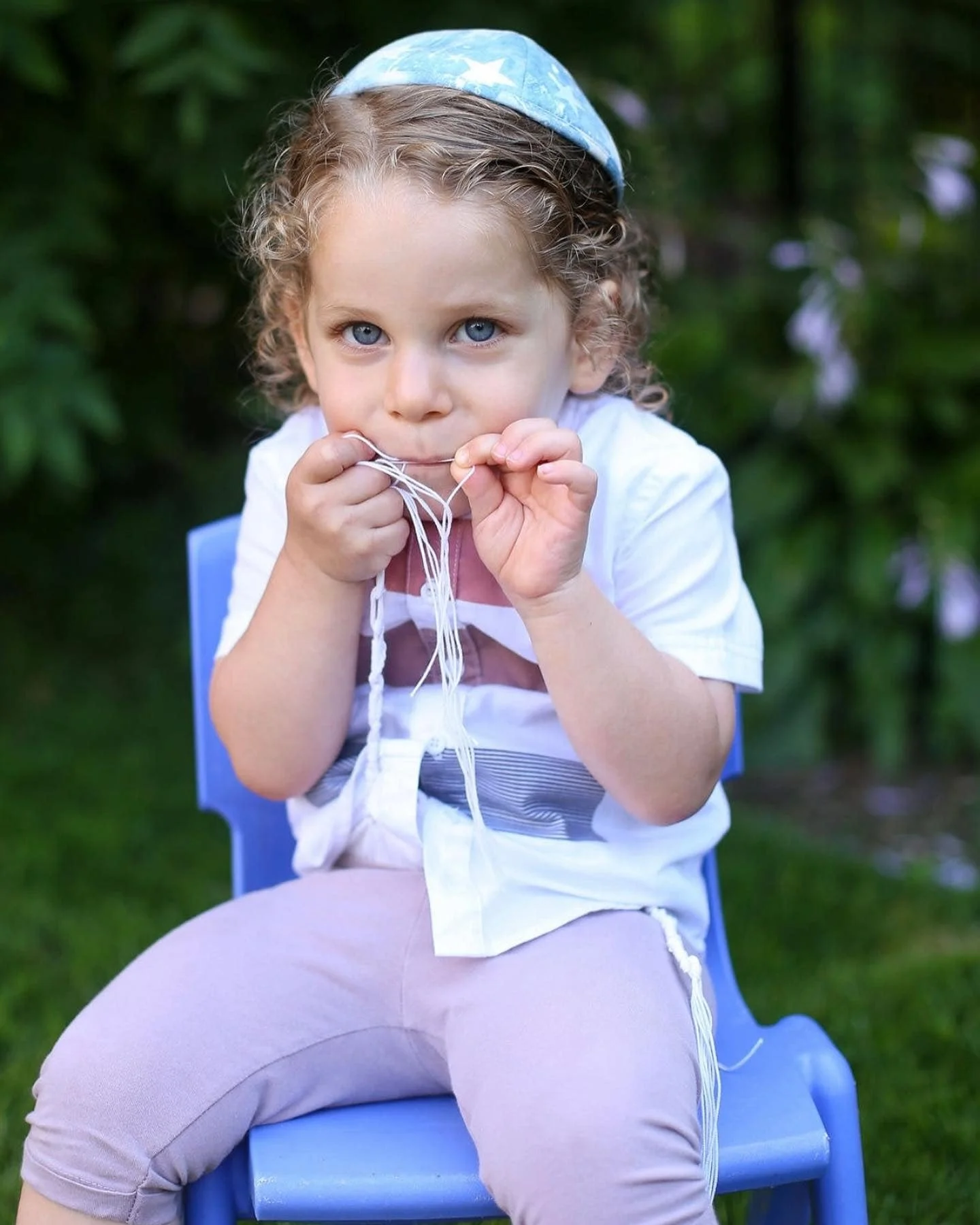 Young child with curly hair and blue eyes sitting on a blue chair outdoors, holding and chewing on a string.