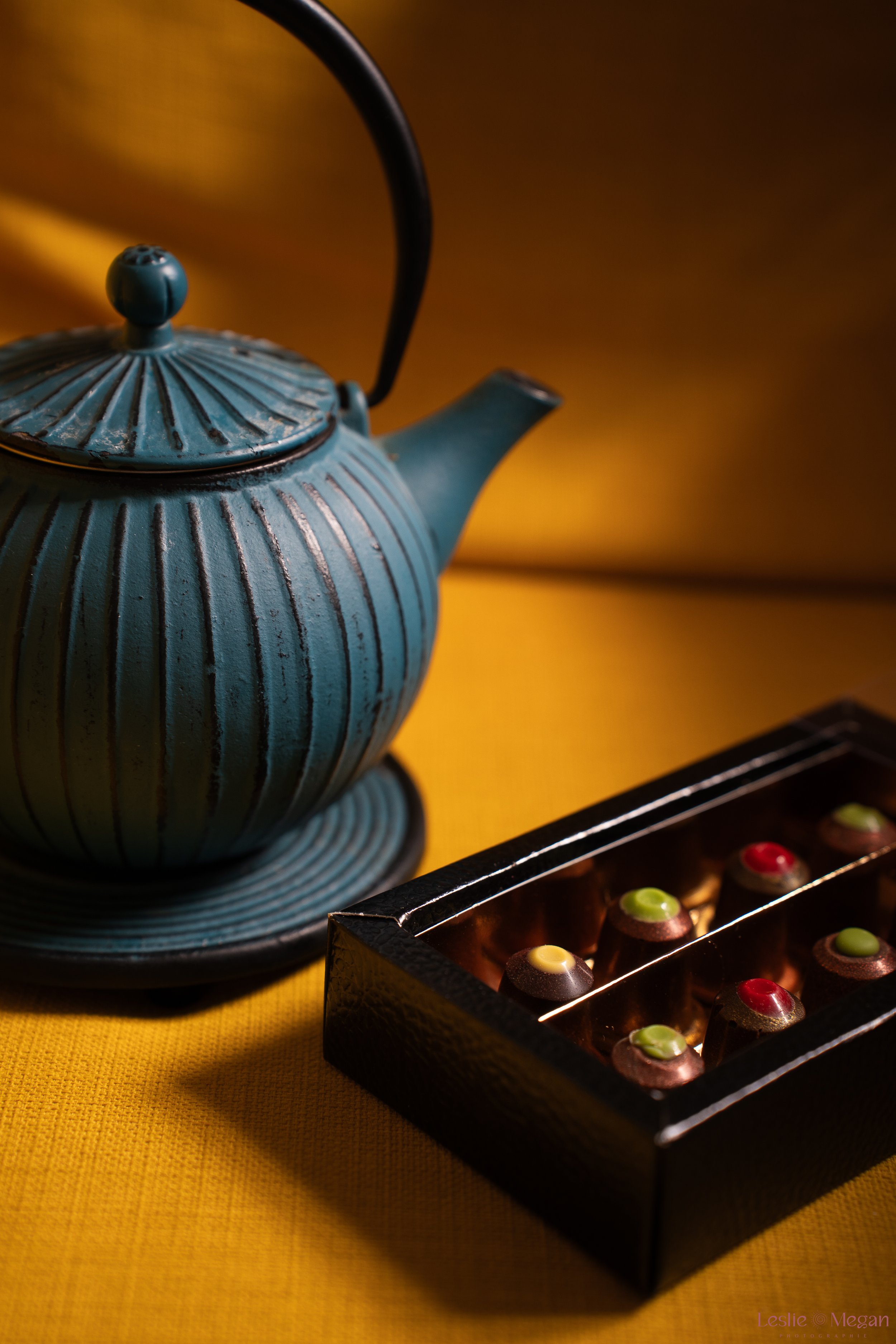 Une théière bleue en métal avec un motif rayé et un bouton à son sommet, placée sur une table jaune avec un plateau de chocolats au thé assortis créés par Carla Campo et Charles Darzacq.