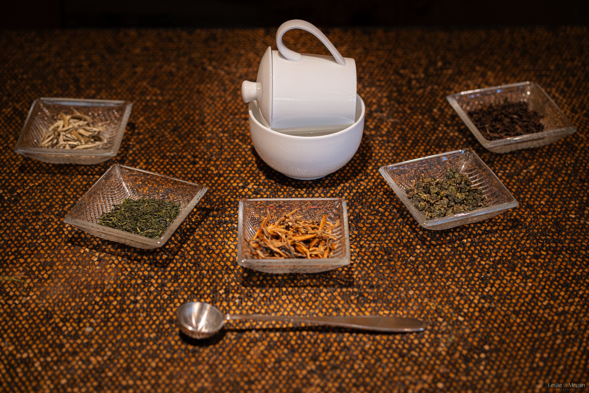 Différents types de thé en feuilles dans des petits bols en verre disposés autour d'une tasse blanche, sur une nappe à motifs géométriques. Une cuillère à mesurer en métal est également présente.