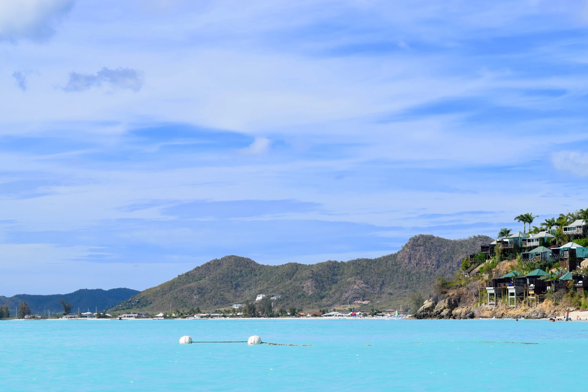 Antigua’s turquoise shoreline framed by gentle hills and hillside villas above the beach — calm water, soft sky, and the island’s relaxed elegance in view.