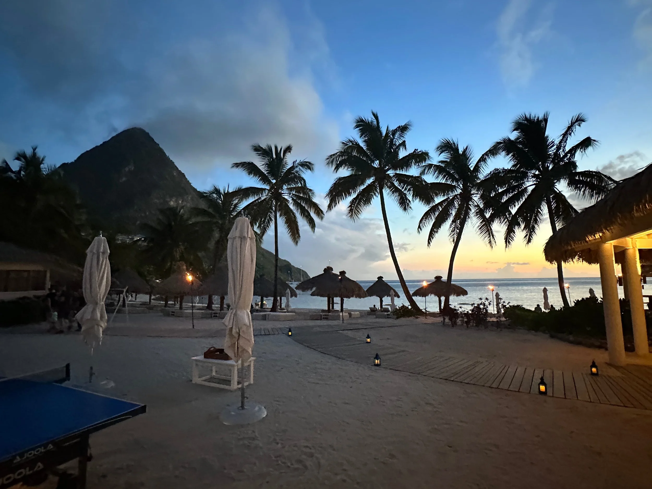St. Lucia beach at dusk with palm trees silhouetted against a blue-and-gold sky, thatched umbrellas on the sand, and calm water beyond.