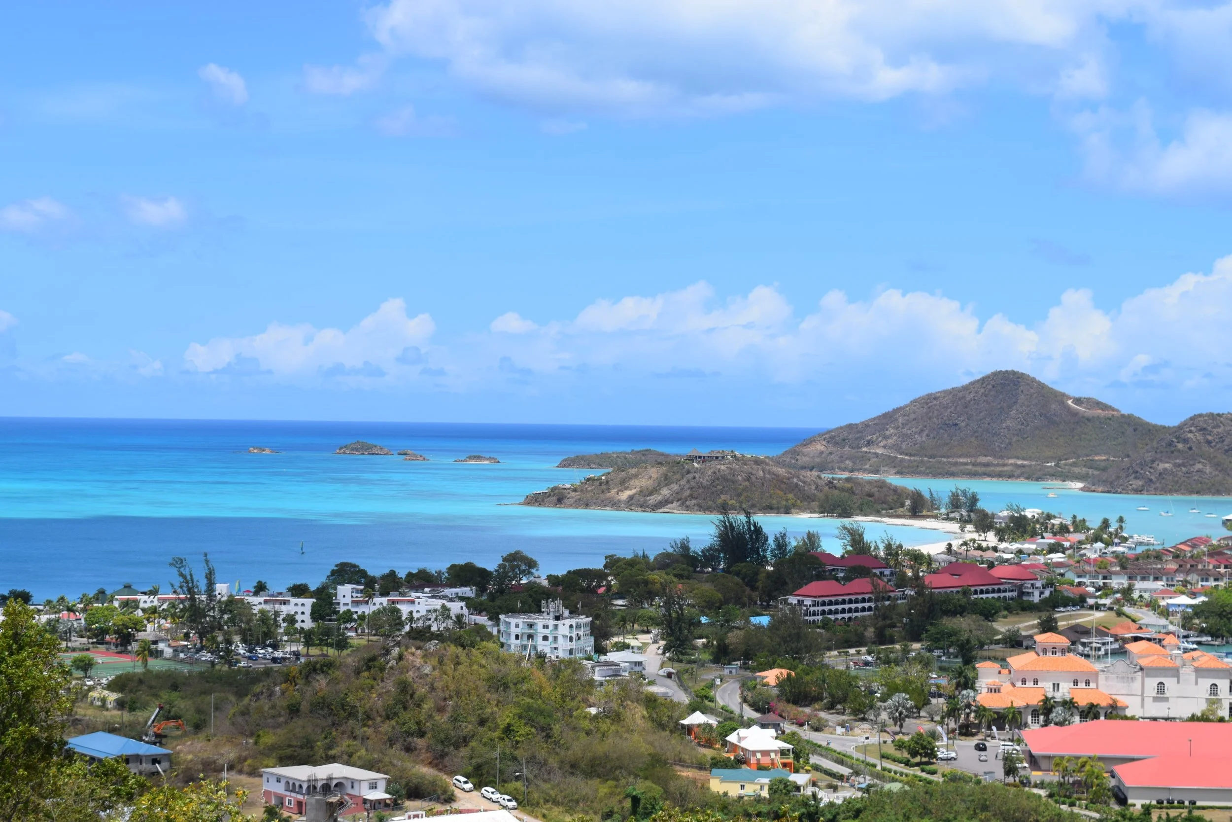 Panoramic view of Antigua’s west coast with red-roofed buildings, calm blue bays, and green hills rolling toward the sea — a colorful snapshot of daily island life.