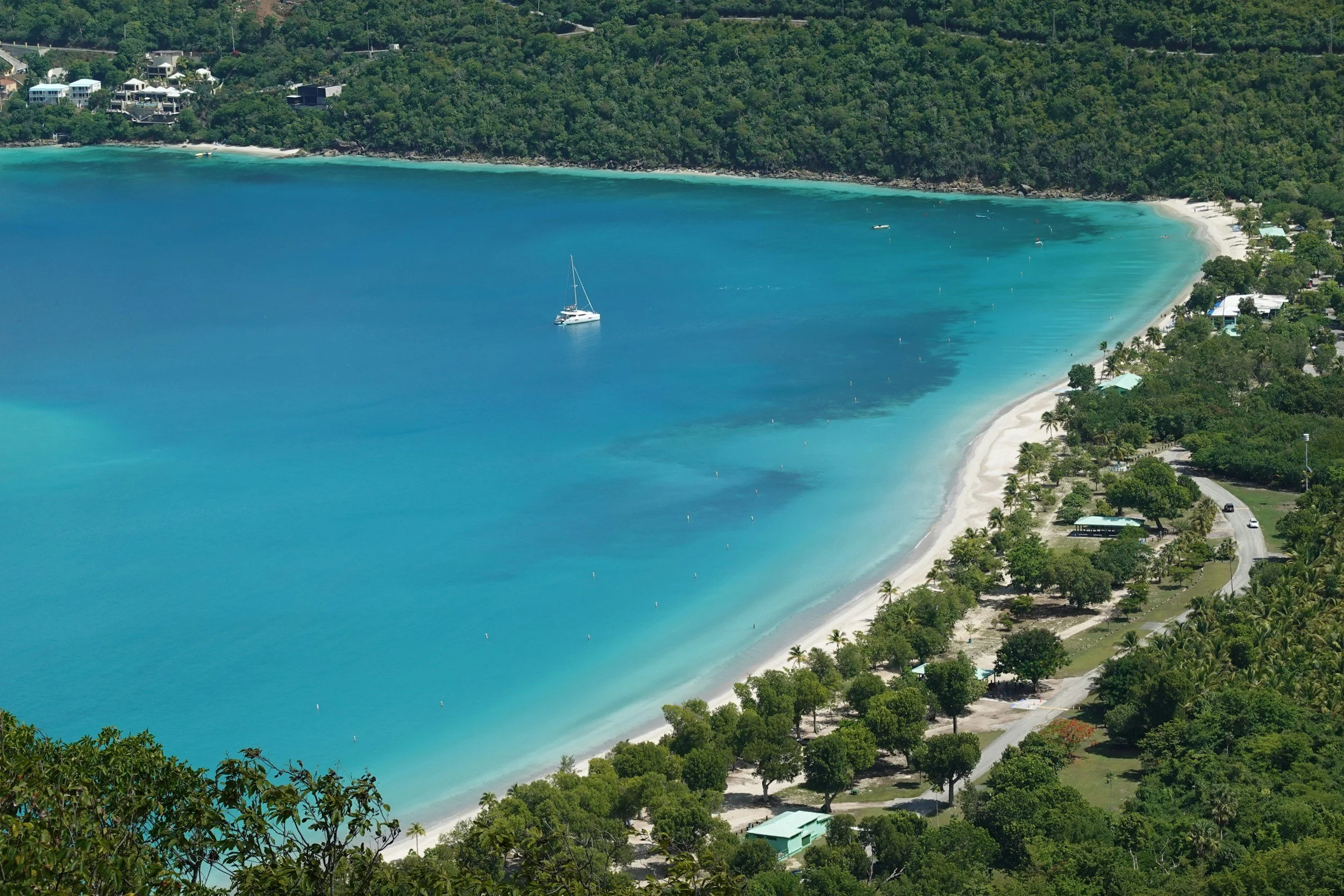 Aerial view of Magens Bay, St. Thomas — long arc of white sand, turquoise water, and palm trees with a sailboat anchored offshore.