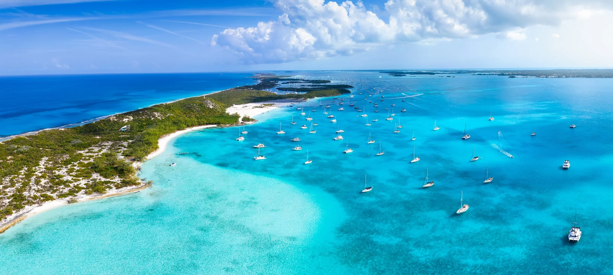 Aerial view of dozens of sailboats anchored in the turquoise shallows of Elizabeth Harbour, Great Exuma, with green cays and sandbars framing the water.