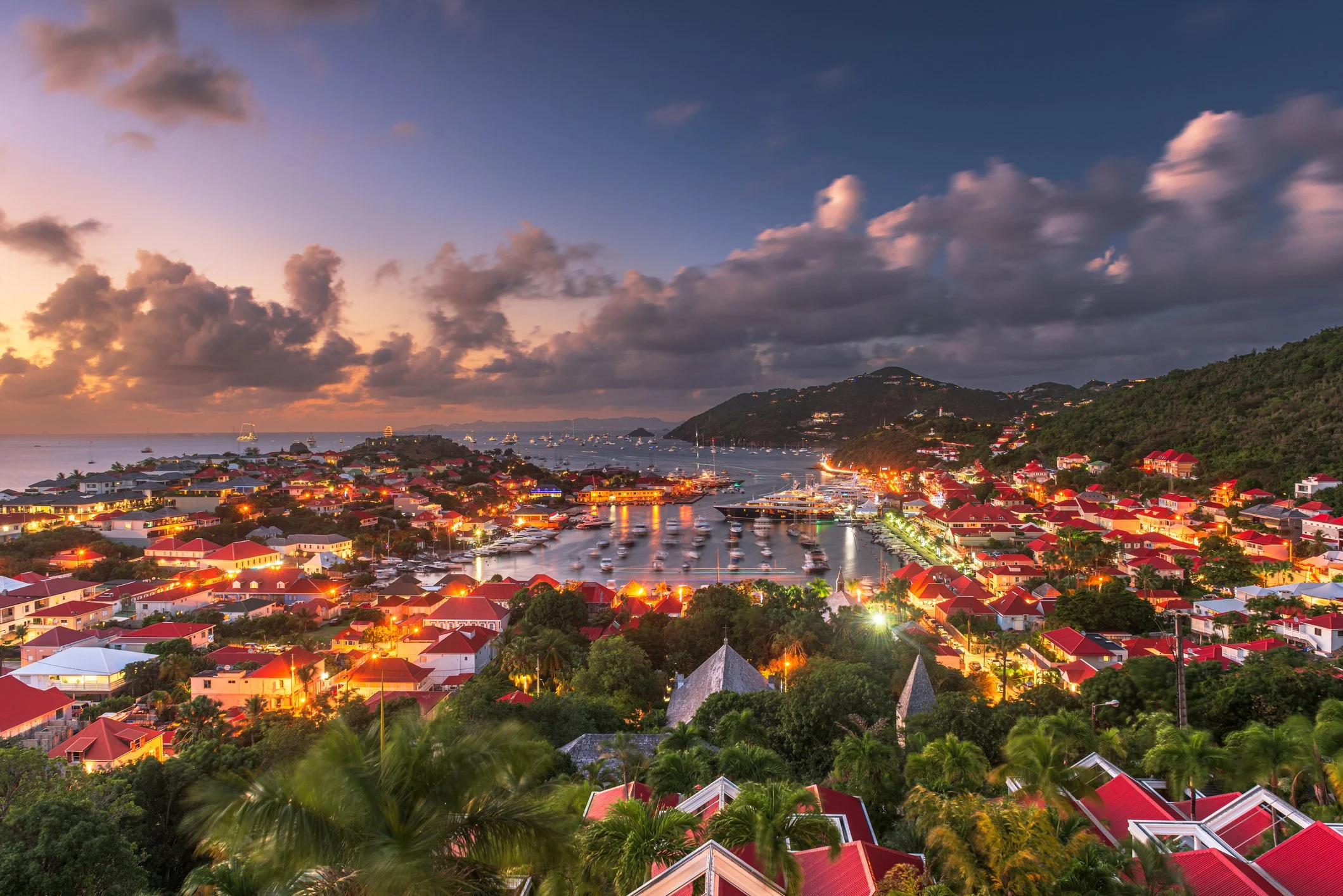 Aerial view of a harbor in St. Barts at night with glowing lights from villas and yachts.