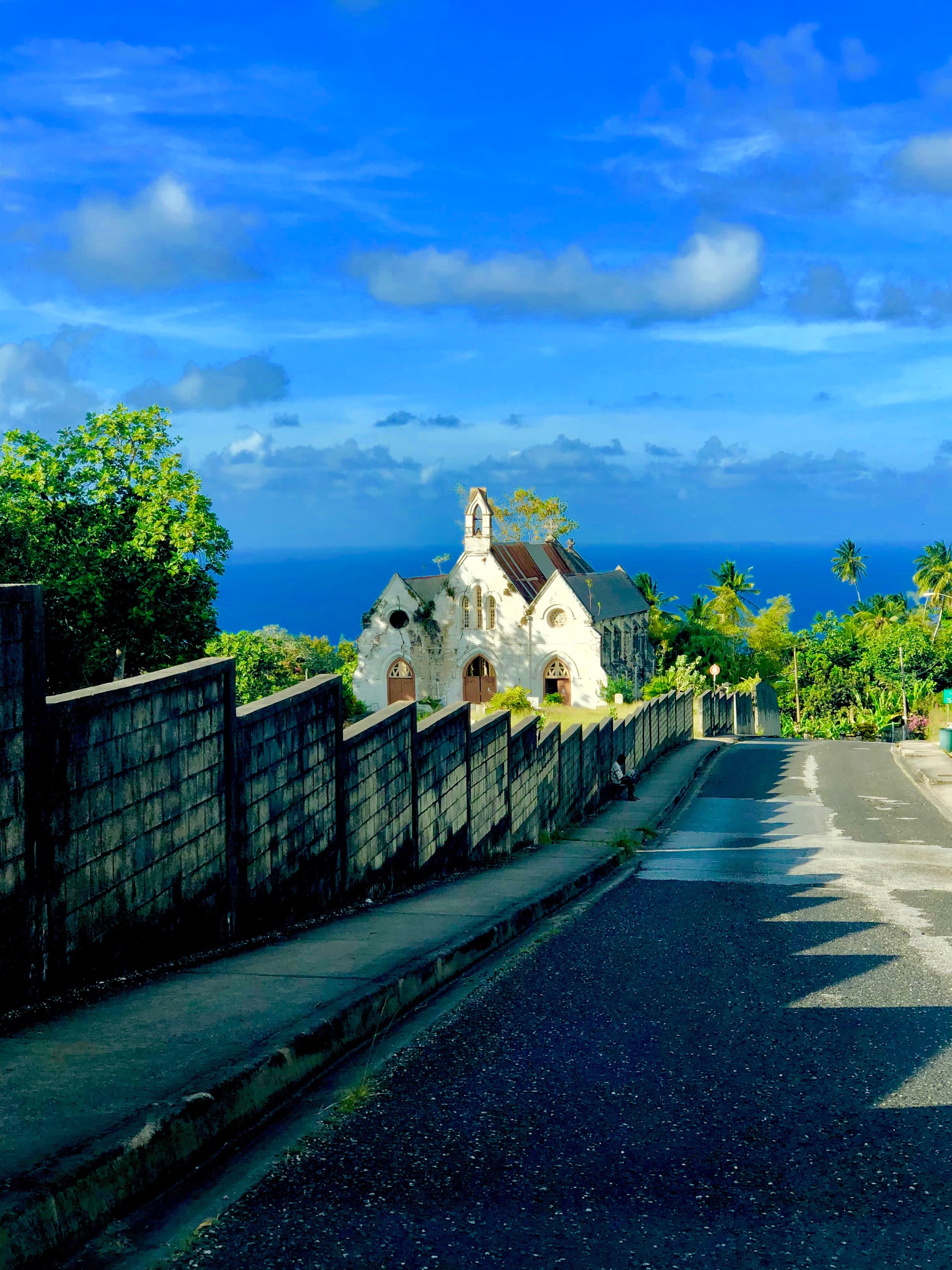 A white stone church on a hill in rural Barbados, framed by palm trees and an ocean backdrop under vivid blue skies.