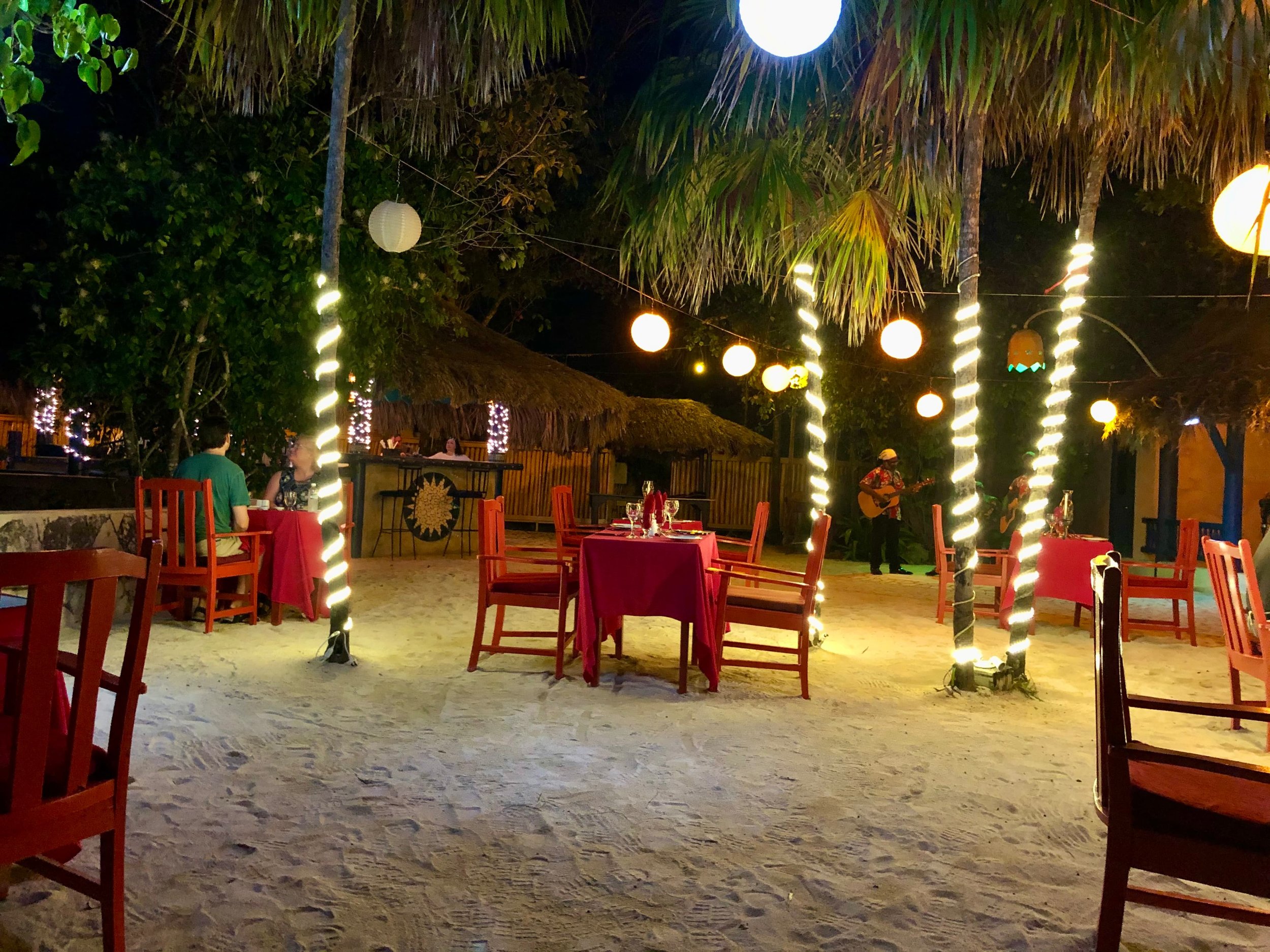Candlelit beach dinner in Jamaica — a table set in the sand under string lights with palm trees and a guitarist playing nearby.