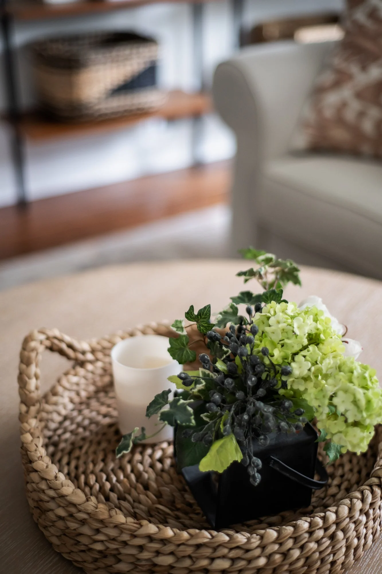 A woven wicker tray with a small white candle and a floral arrangement of green hydrangeas, dark berries, and green leaves on a beige surface in a cozy living room.