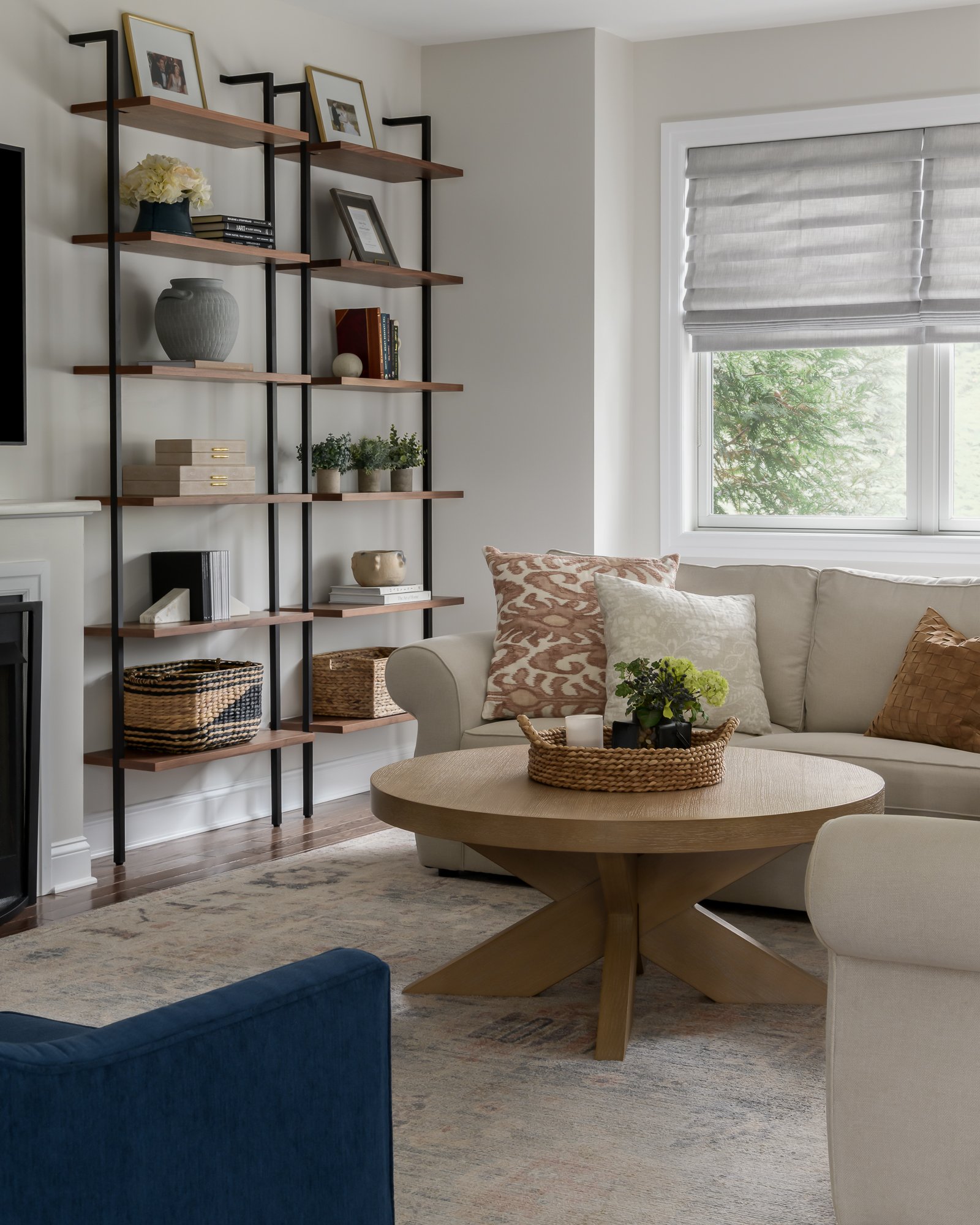 Living room with a white sofa, patterned pillows, a round wooden coffee table with a woven tray and candles, a window with gray blinds, a tall shelf with books and decorative items, and a blue armchair.