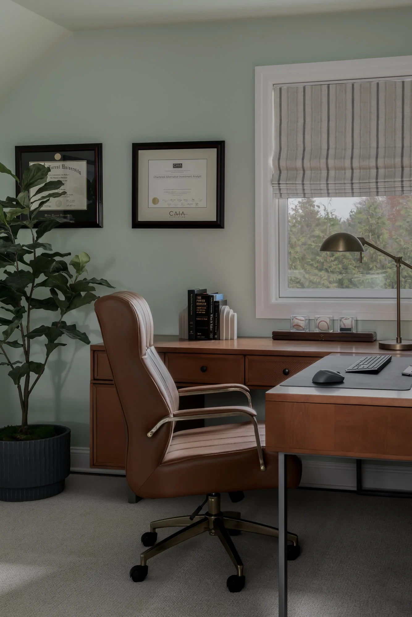 Home office with wooden desk, tan leather office chair, framed diplomas on light green wall, window with a striped valance, potted plant, books, and desk lamp.