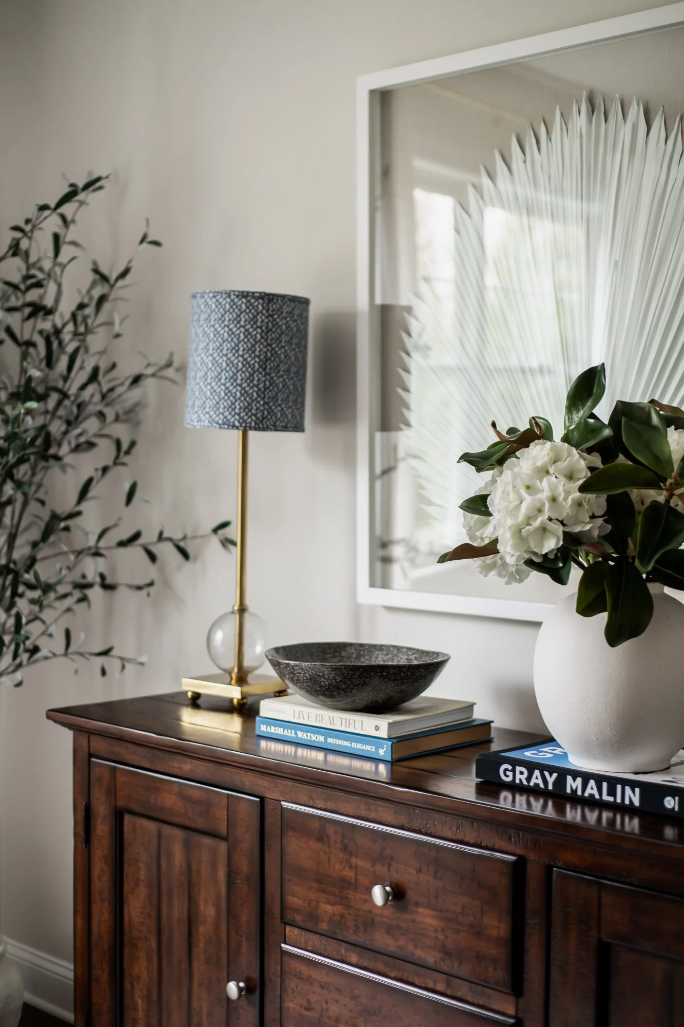A wooden sideboard with a black ceramic bowl, books, and a vase of white flowers, beside a large mirror and a table lamp.