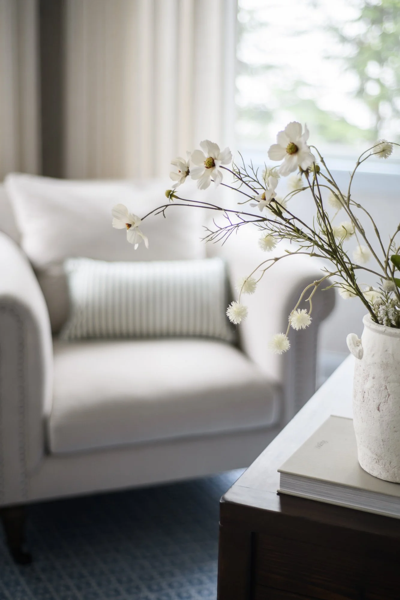 A white ceramic vase with white flowers on a dark wooden table in a cozy, light-filled living room with a white sofa and striped pillows.