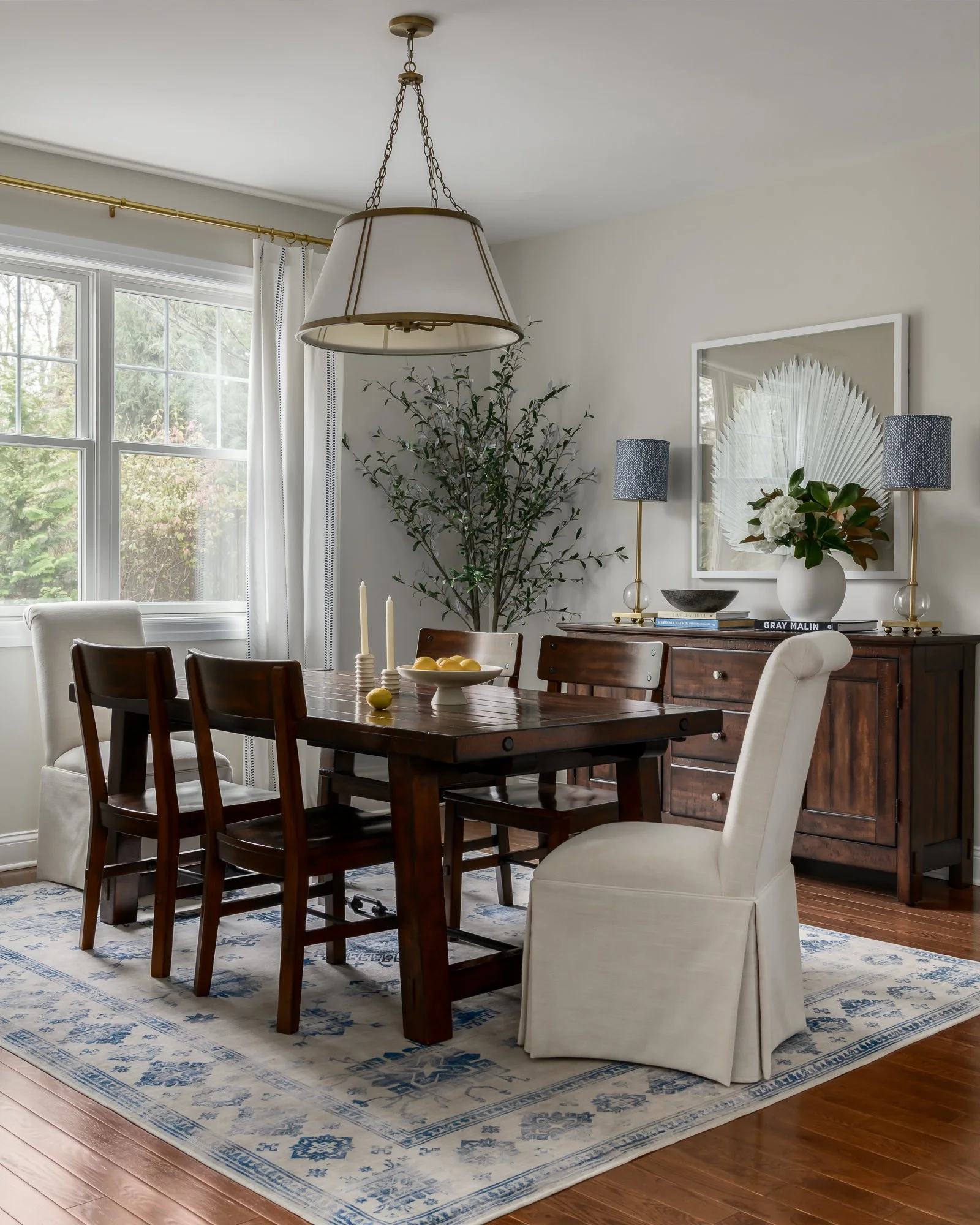 A breakfast or dining room with a dark wood table and six chairs, two white and four dark wood. A large window with white curtains is on one side, letting in natural light. A white and blue patterned rug is beneath the table. In the background, a woo