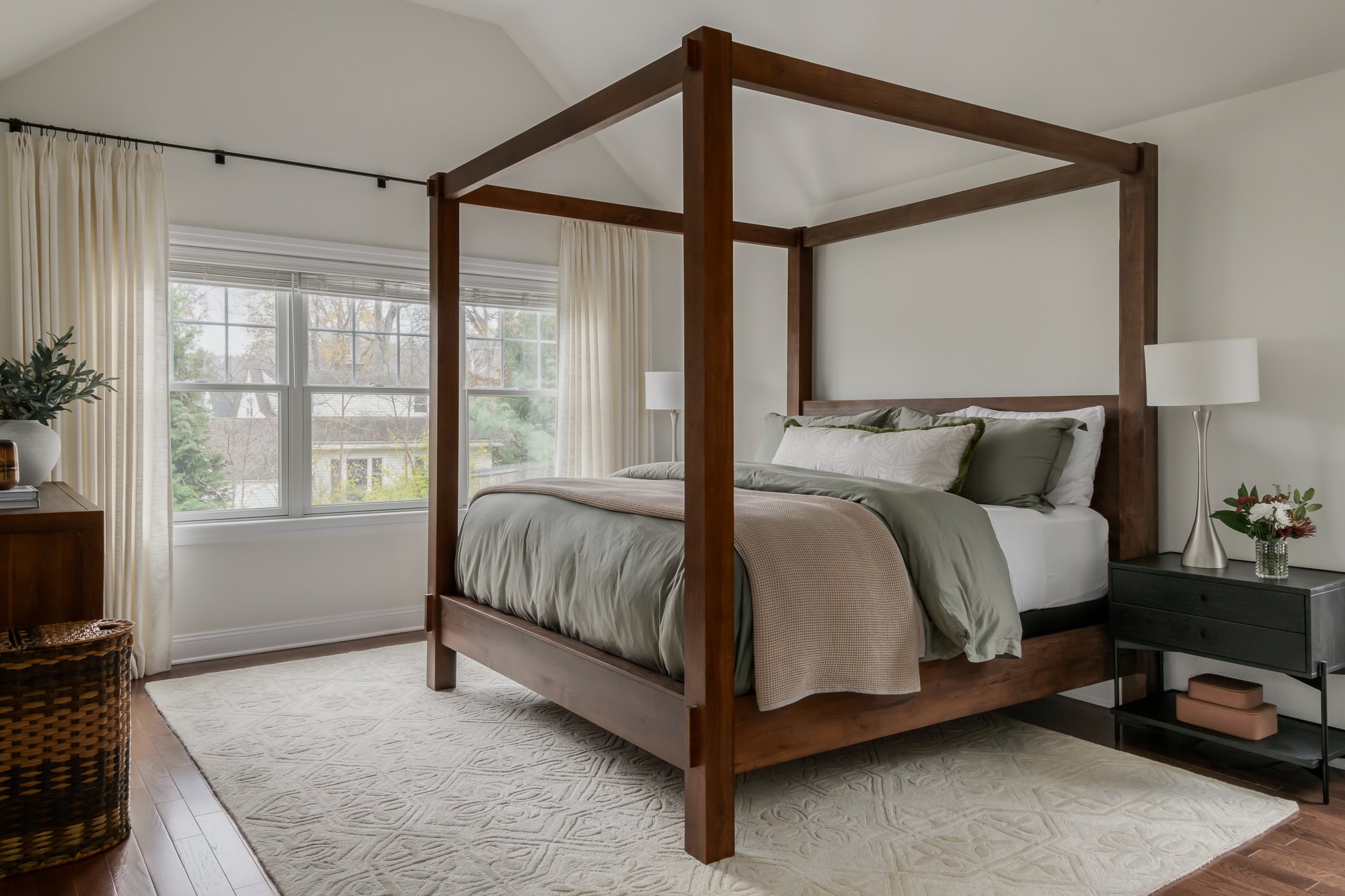 A bedroom with a wooden four-poster bed, light green and white bedding, a black nightstand with a lamp and flower vase, large window with cream curtains, hardwood floors, and a cream area rug.