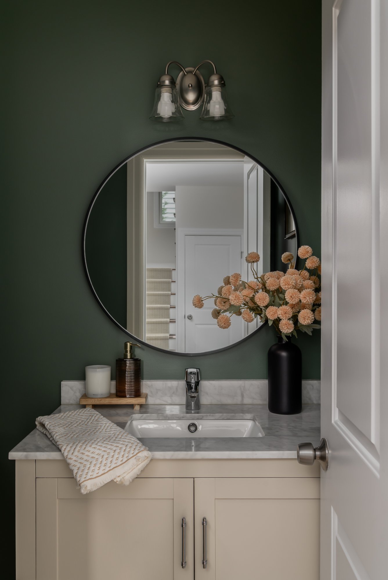 Bathroom vanity with beige cabinet, marble countertop, oval mirror, black vase with pink flowers, white candle, soap dispenser, towel, and light fixture above mirror.