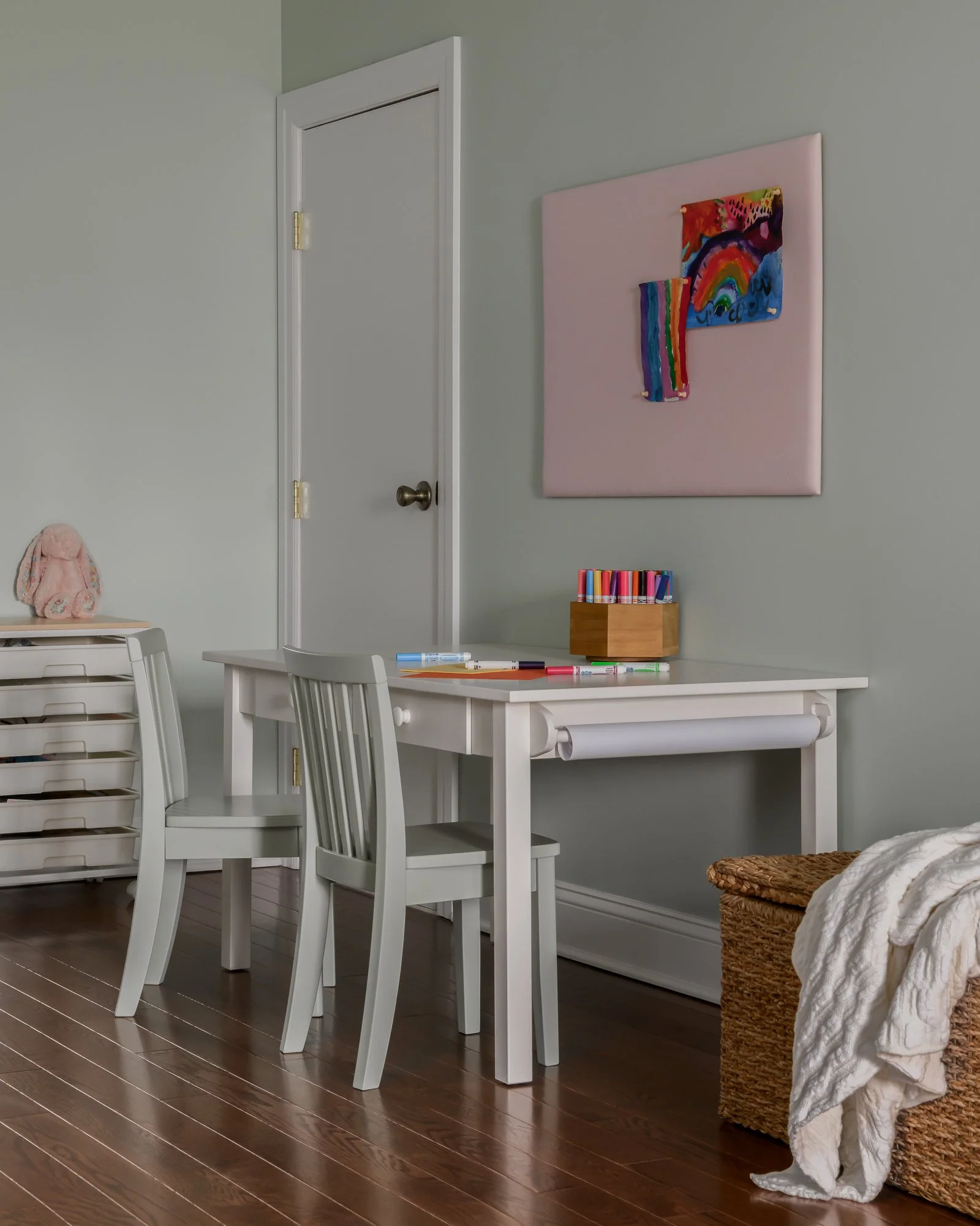 A small white table with two white chairs, a wooden box with colorful markers, and a pink abstract rainbow artwork on a light green wall in a child's room. A wicker basket with a blanket is nearby.