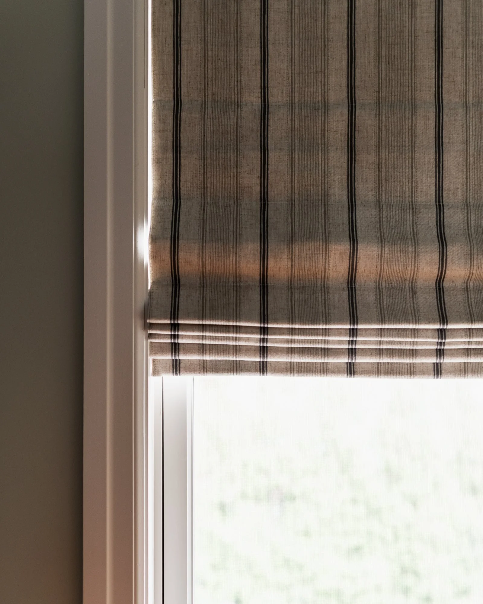 A window with a beige and black striped roman shade partially drawn, allowing light to filter through.