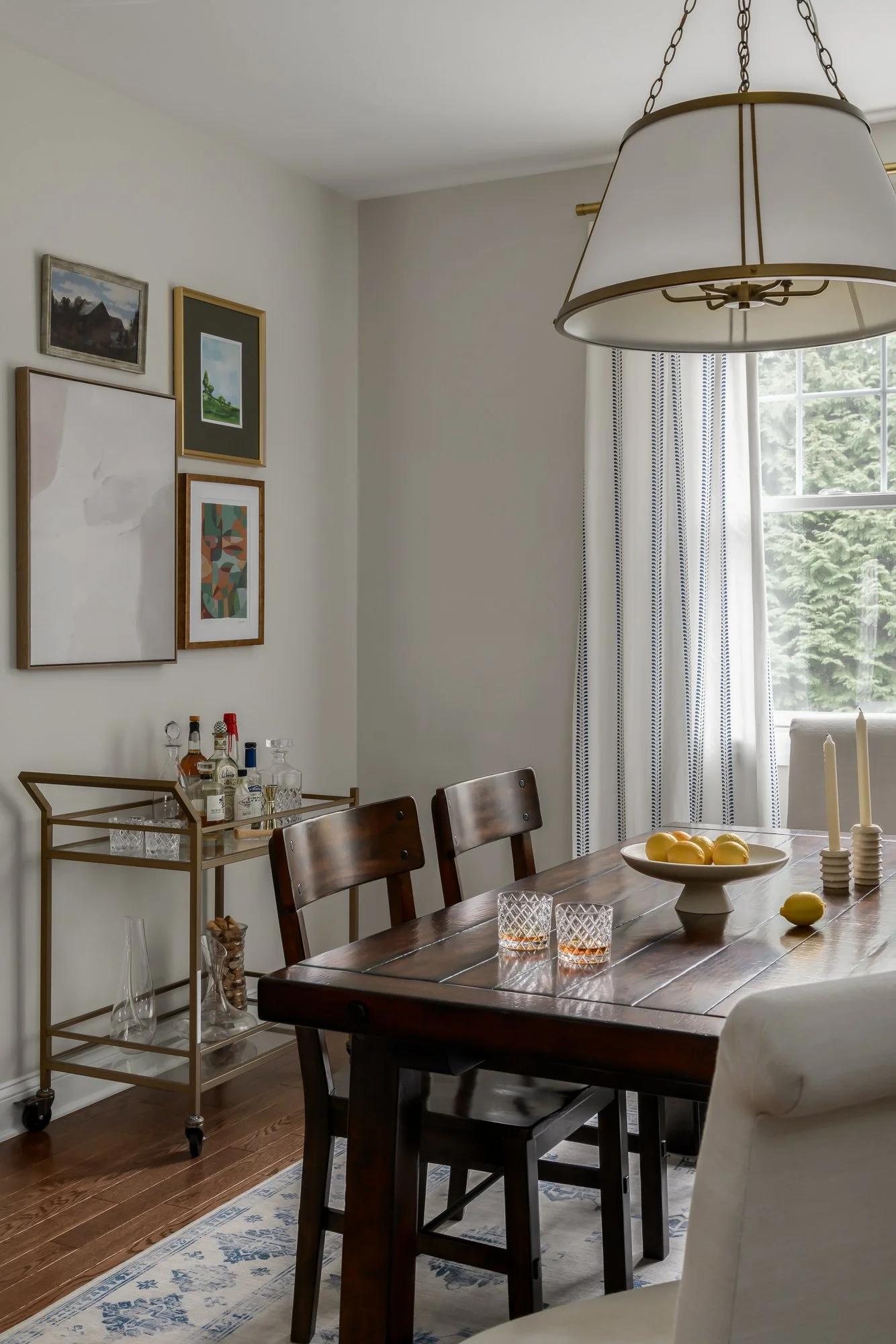 A dining room with a wooden table, four chairs, a light fixture hanging above, and various decorative items including a bowl of lemons, candles, and glassware. There is a window with curtains, and a wall with art paintings. A bar cart with bottles an