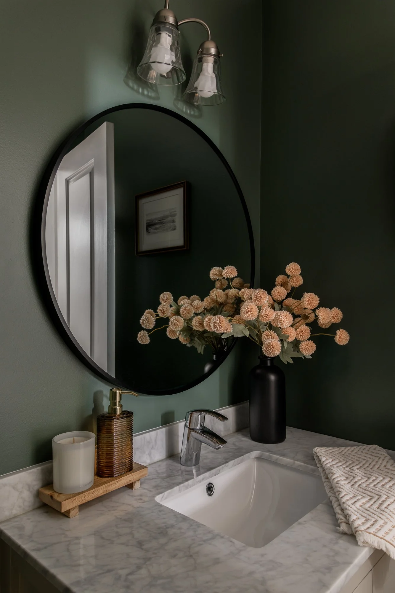 Bathroom vanity with marble countertop, black vase with pink flowers, mirror, wall light fixture, soap dispenser, candle, and folded towel.