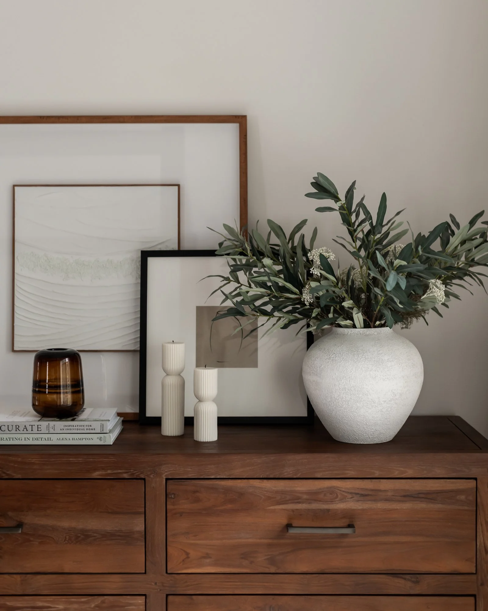 Decorative interior with framed artwork, a large white vase with greenery, two white textured candles, a small brown glass vase, and books on a wooden sideboard.