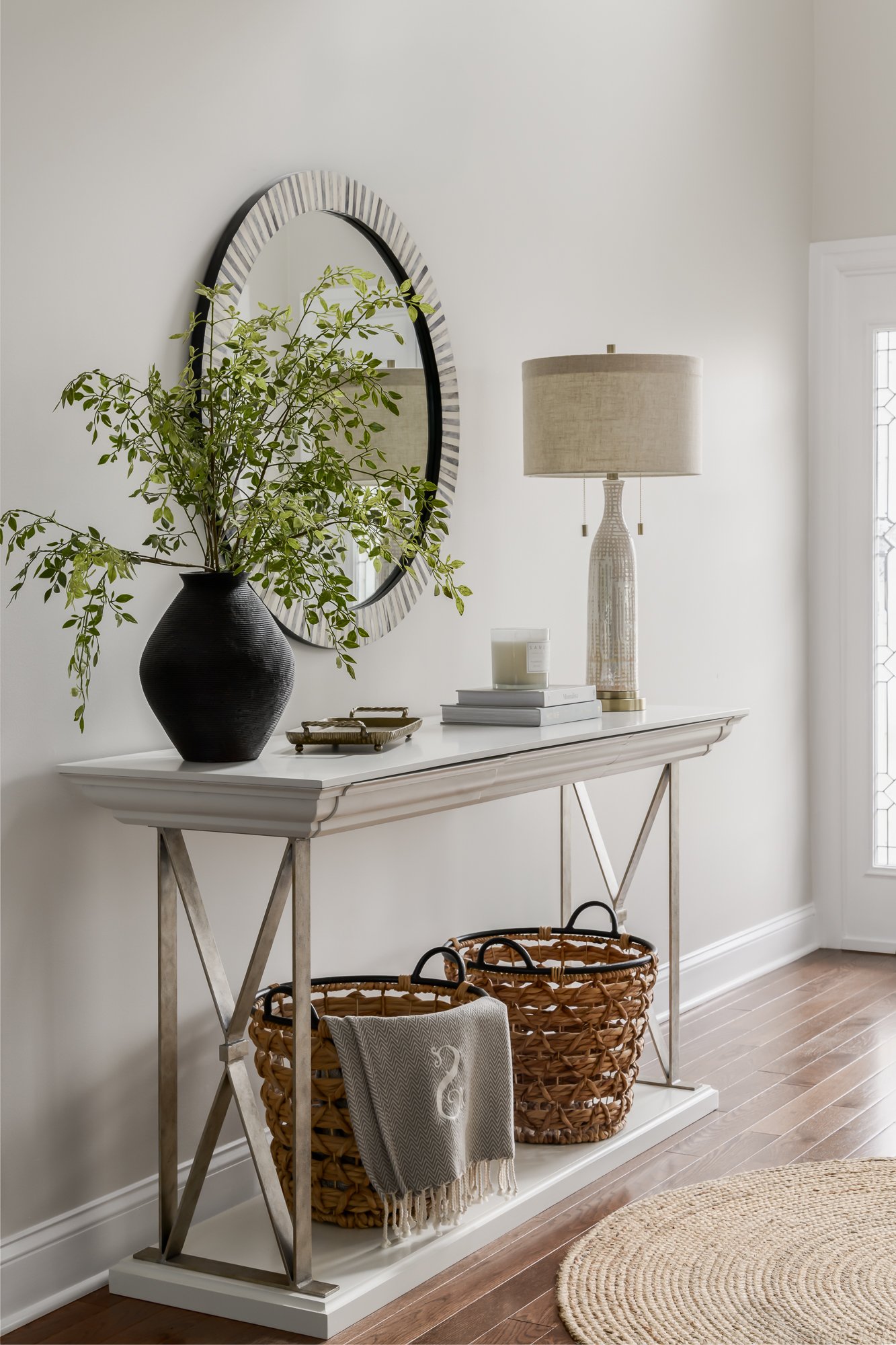 A white hallway console table decorated with a black vase containing greenery, a lamp with a beige shade, and a stack of books with candles on top. Two wicker baskets are placed underneath, and a decorative throw blanket hangs over one basket. A larg