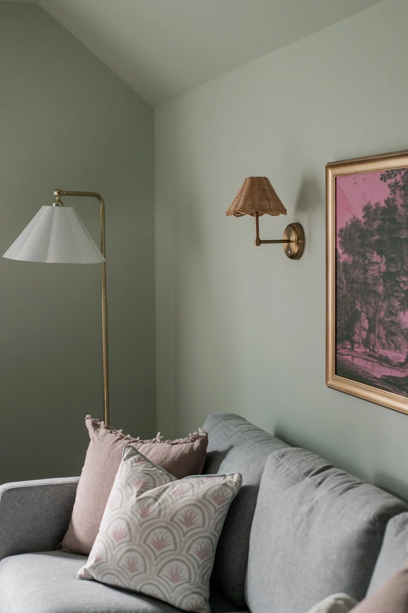 A living room with a gray sofa adorned with beige and patterned pillows, a standing lamp with a white shade, a wall-mounted wicker lampshade with a brass arm, and a framed landscape picture on a light green wall.
