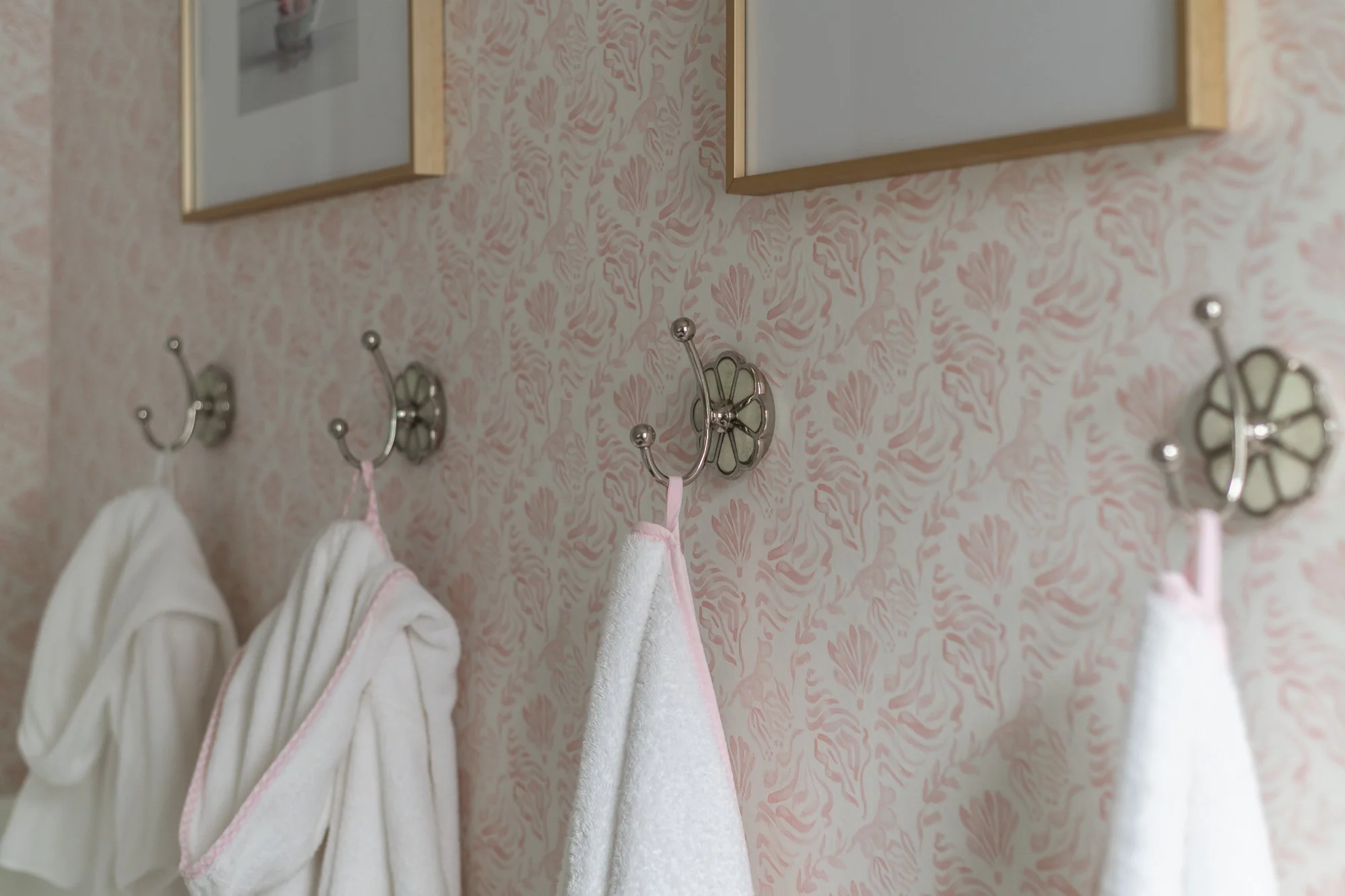 Close-up of vintage-style bathroom hooks with hanging towels on a pink floral wallpapered wall, above framed pictures.