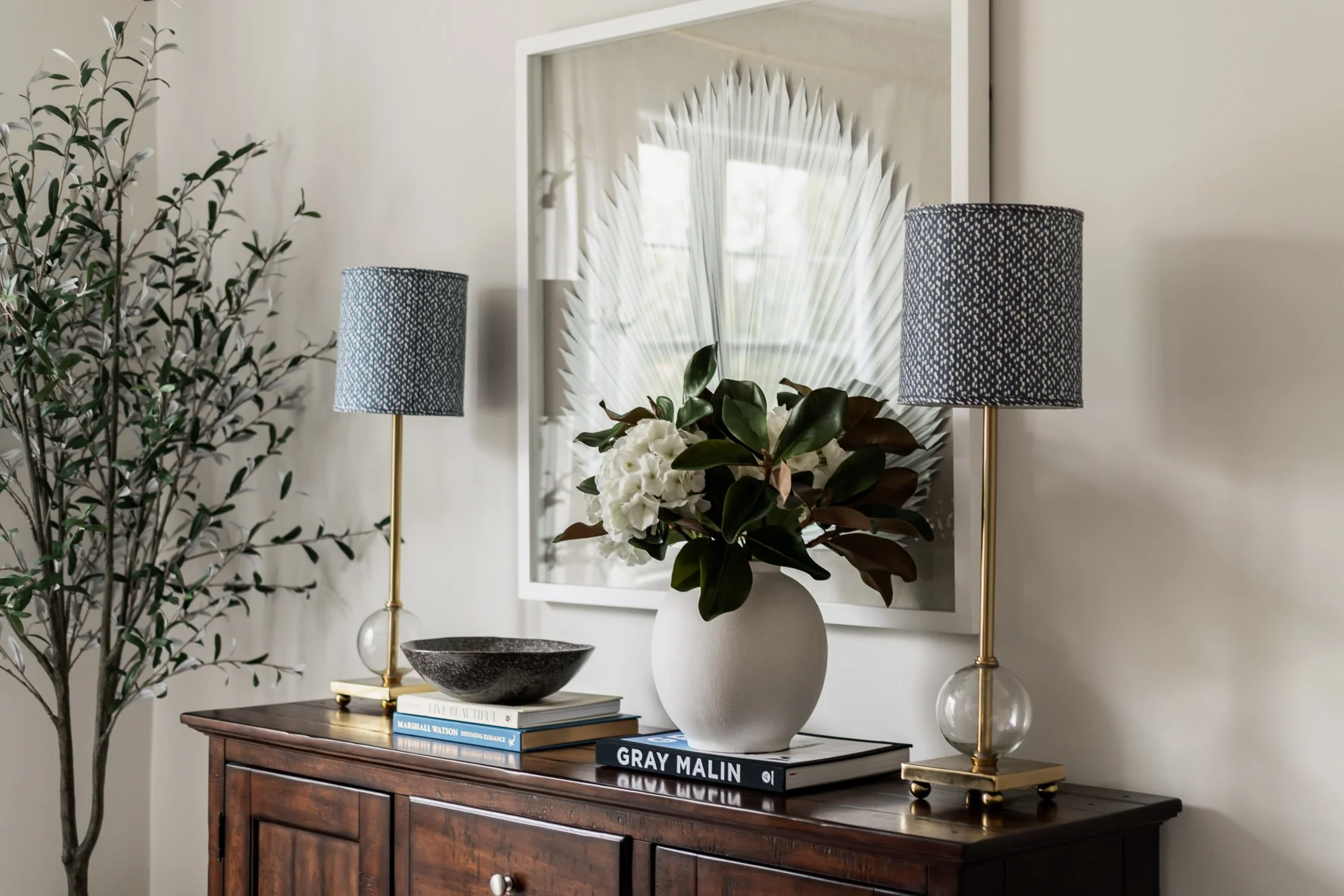 Decorative console table with a vase of white flowers, two black and white lamps, a bowl, and books, with a large framed wall art piece behind.