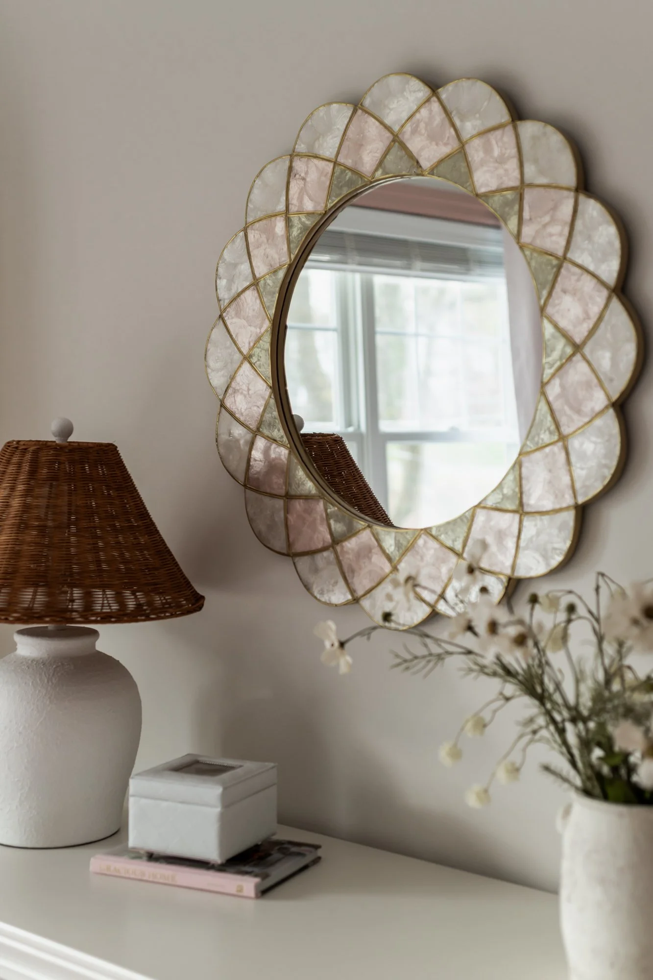 Decorative wall mirror with a scalloped frame made of pink and white marble segments, a wicker table lamp, a white box, a stack of magazines, and a flower vase with white blossoms on a white dresser.