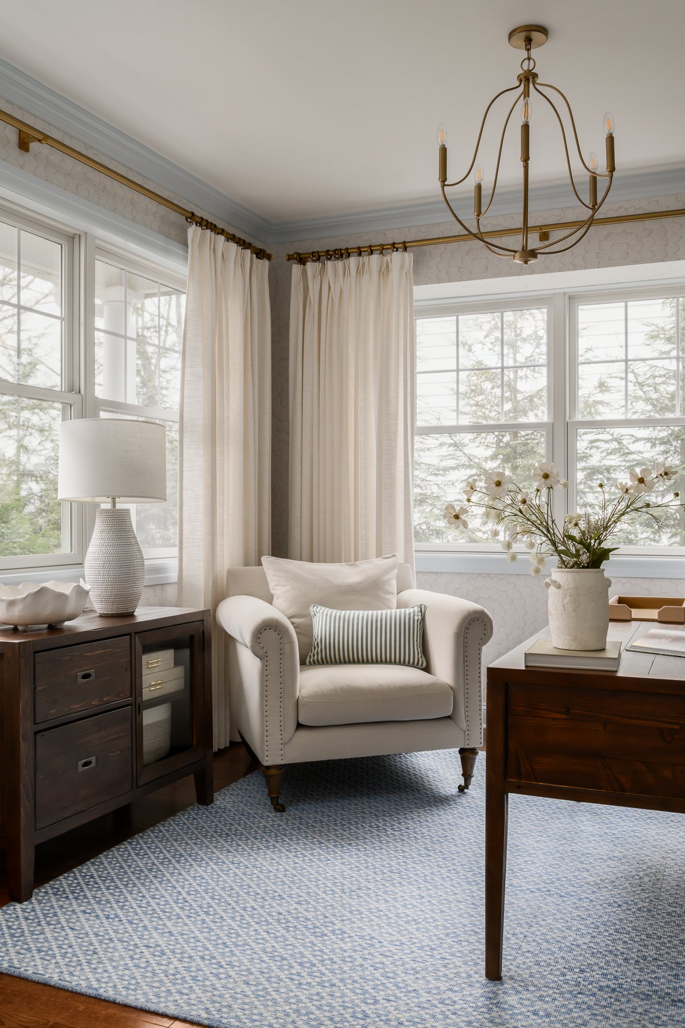 A cozy living room corner with large windows, cream curtains, a white armchair with pillow, a wood sideboard with a table lamp and bowl, a vase of flowers, and a blue patterned rug.