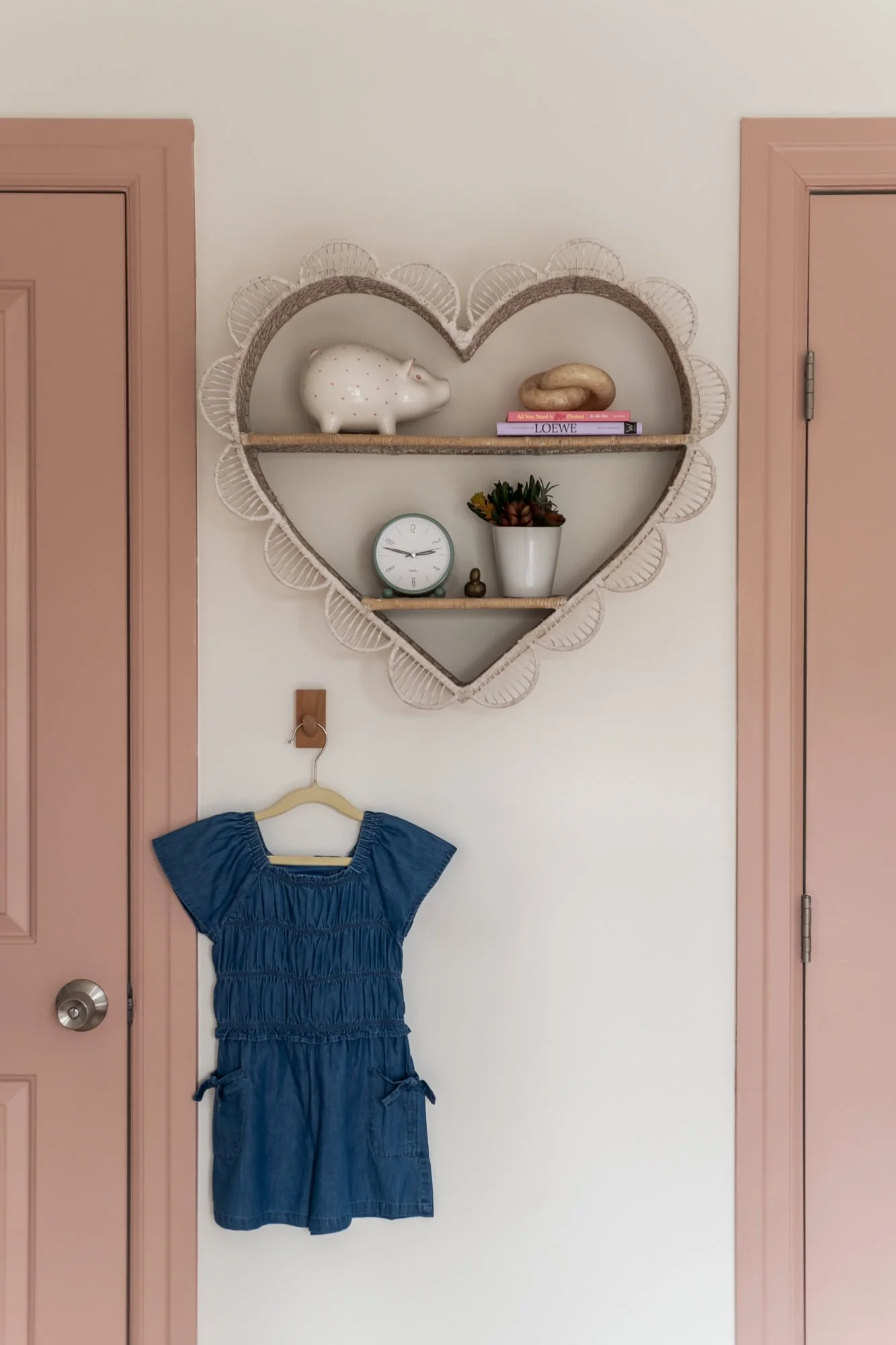 A wall with a pink door on each side, a hanging blue dress, and a heart-shaped shelf with decorative items including a pig figurine, a book, a clock, a plant, and a small figurine.