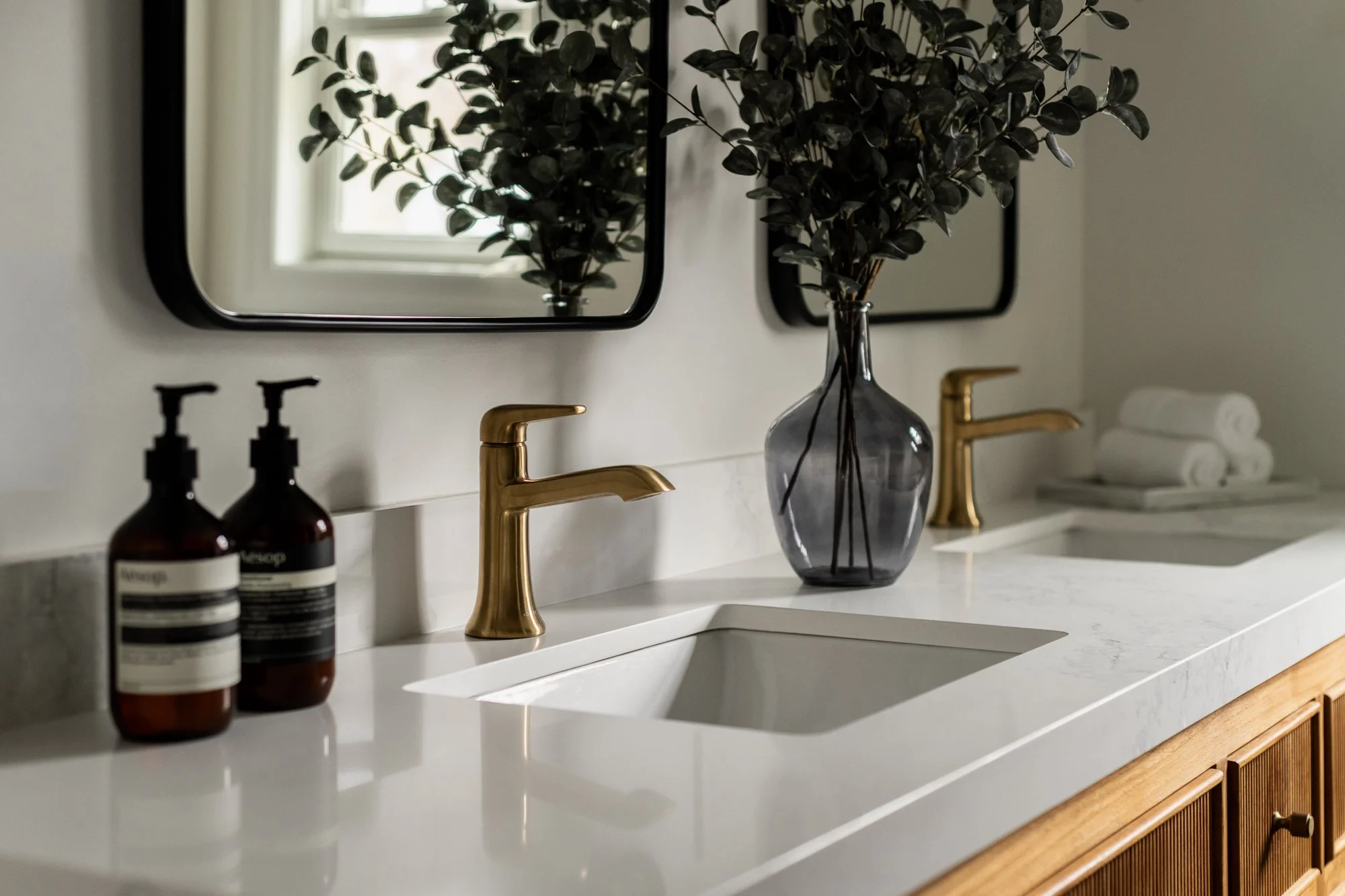 Bathroom vanity with a white countertop, two gold faucets, a large glass vase with greenery, two mirrors, bottles of soap, and rolled towels in the background.