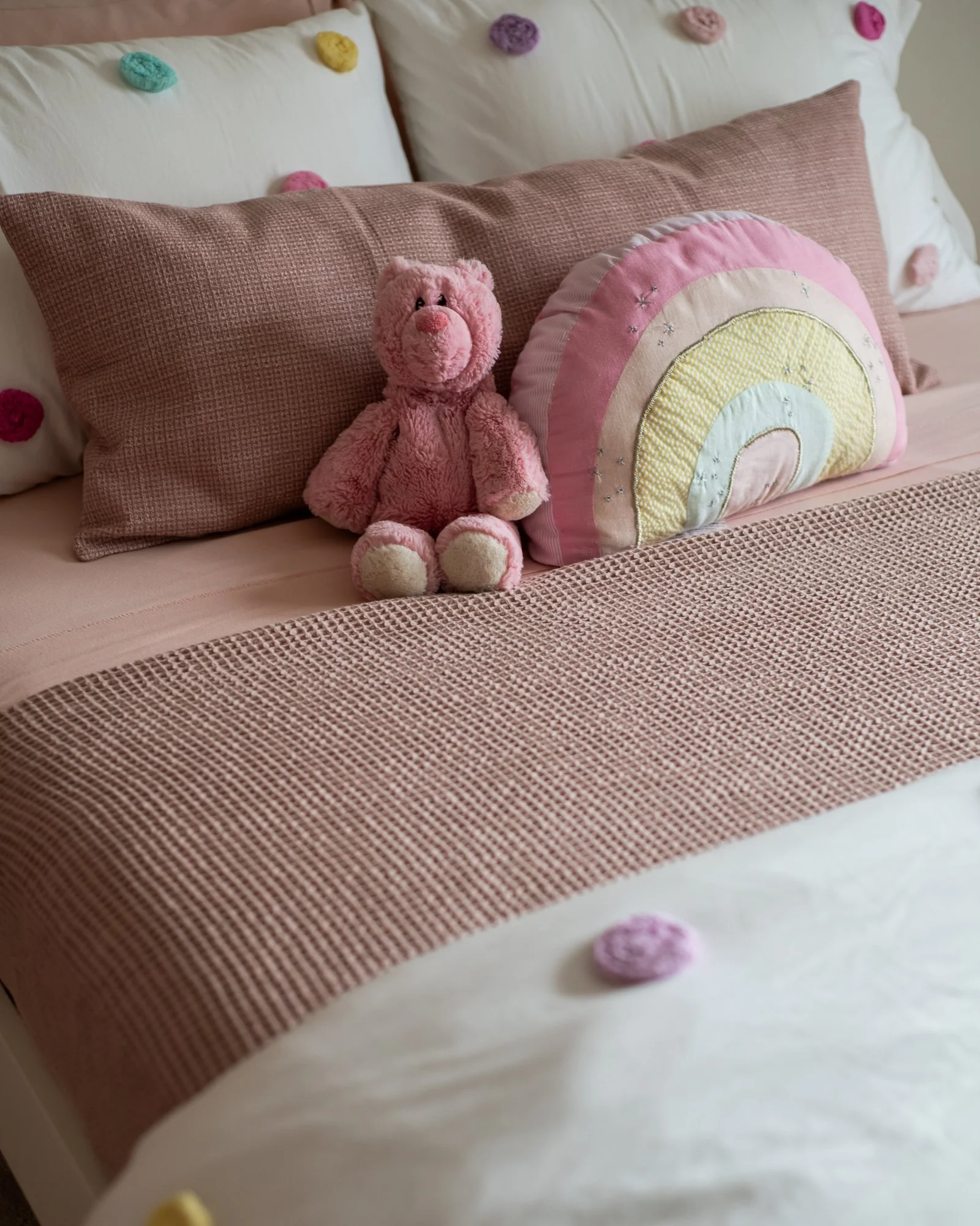 A neatly made bed with pink and white bedding, decorated with a pink teddy bear, a rainbow-shaped pillow, and colorful pom-pom pillows on the headboard.